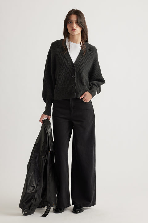 Woman wearing a charcoal knit cardigan with balloon sleeves, paired with wide-leg black pants and holding a leather jacket.
