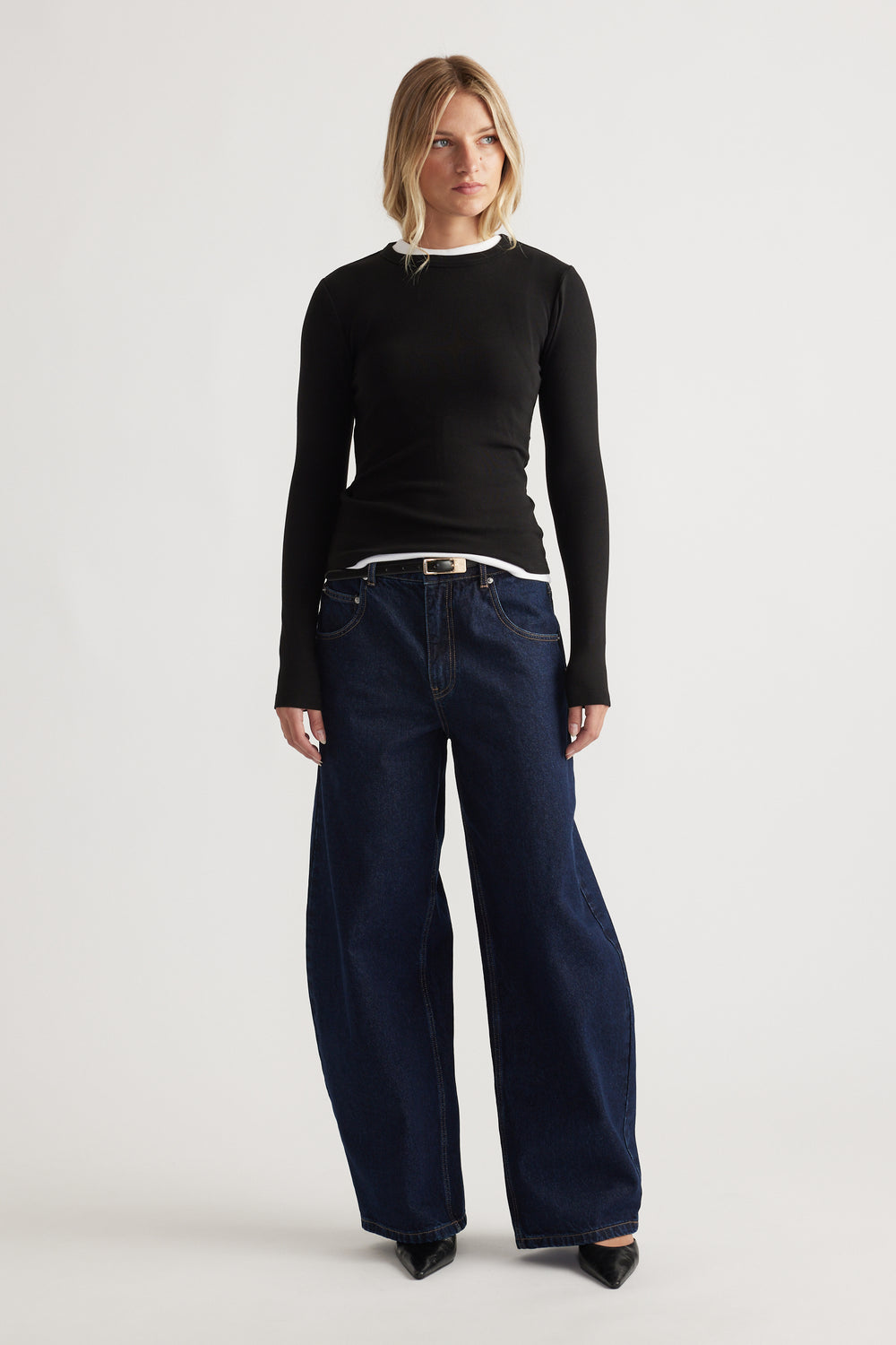 Woman wearing a fitted black long sleeve top layered over a white shirt with wide-leg dark blue denim jeans and black heels