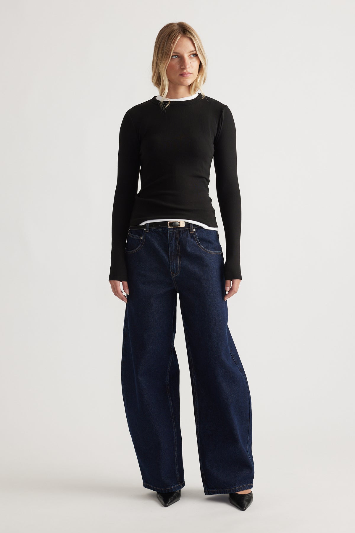 Woman wearing a fitted black long sleeve top layered over a white shirt with wide-leg dark blue denim jeans and black heels