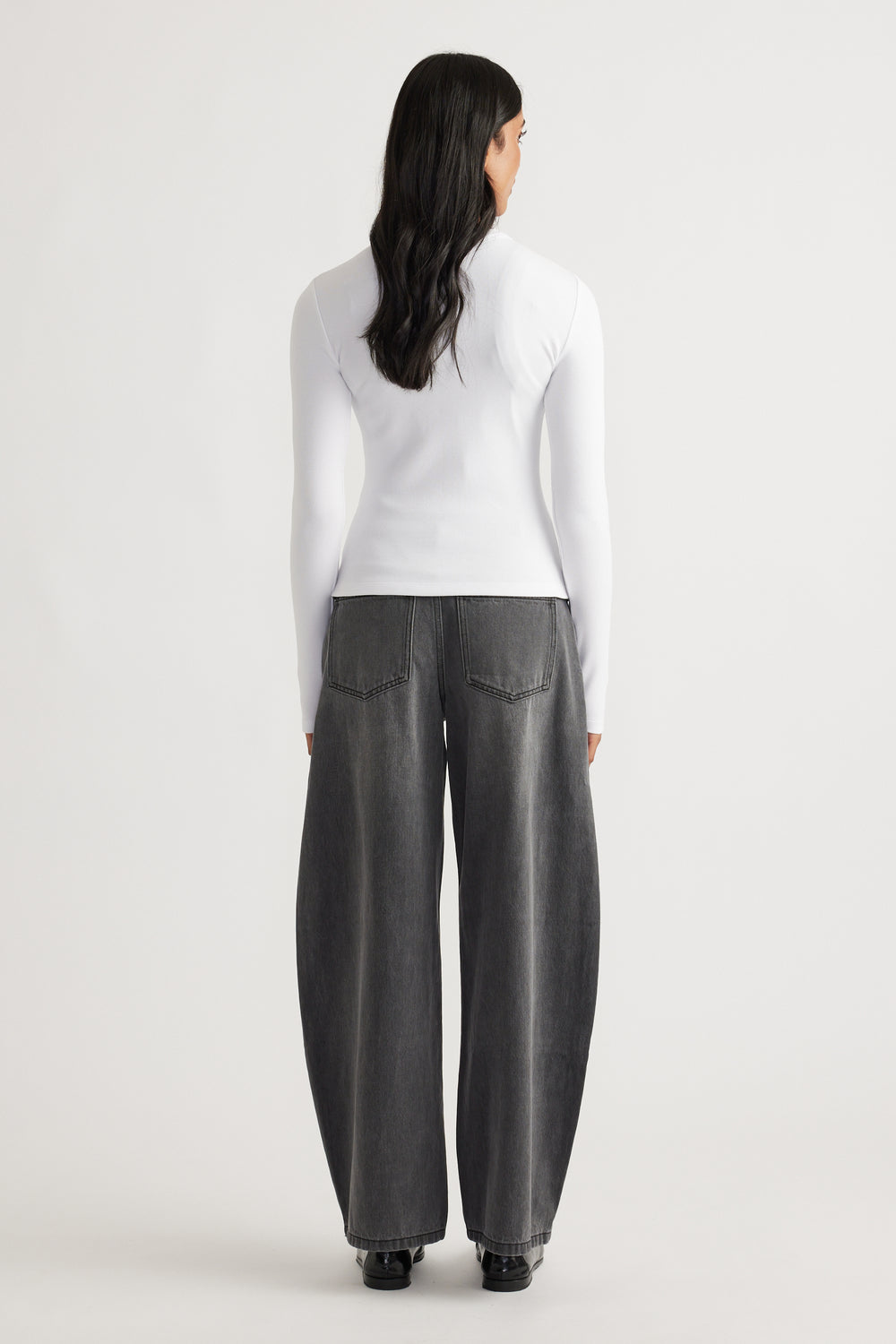 Woman wearing a fitted white long sleeve top paired with wide leg faded black denim jeans and black shoes viewed from behind