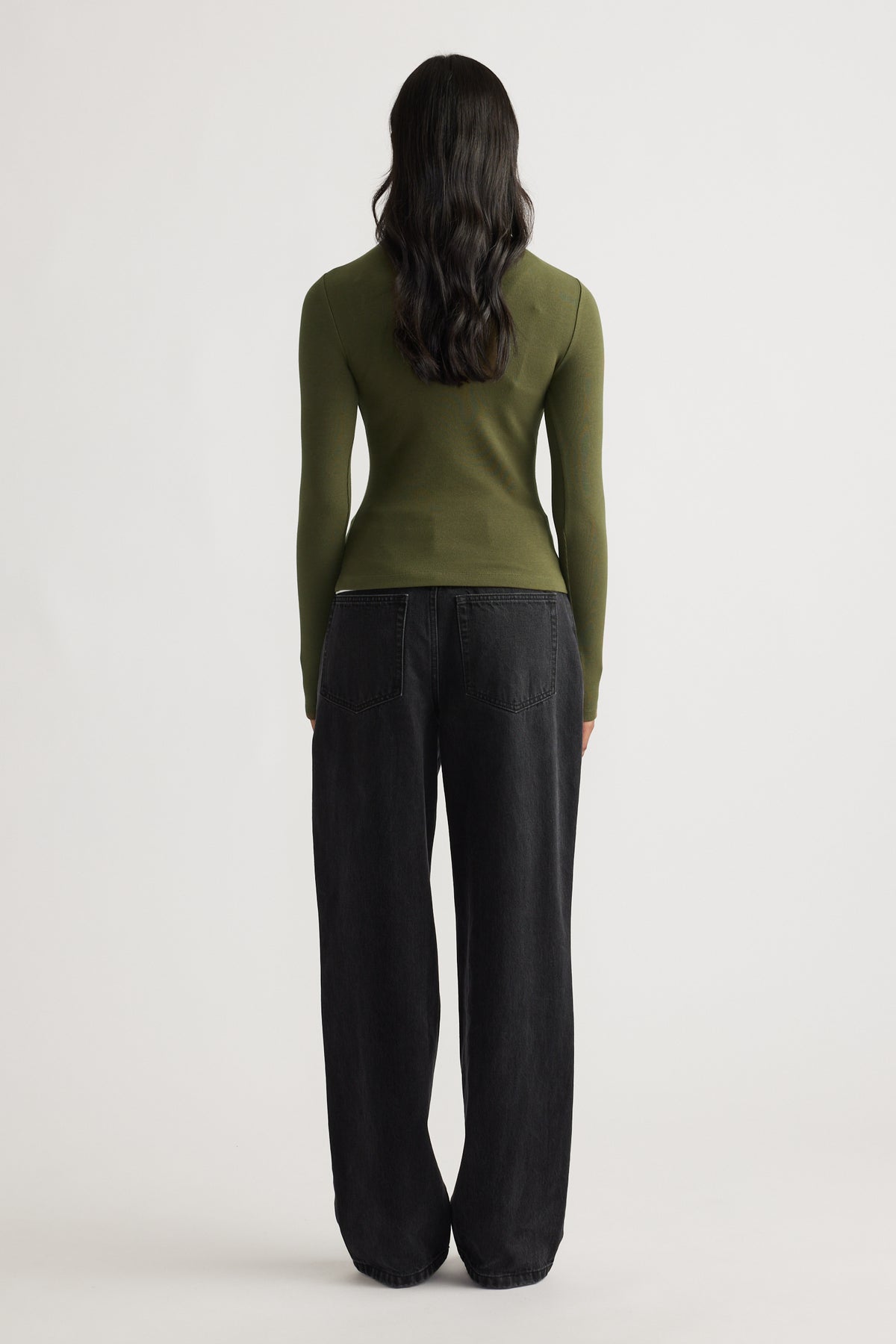 Woman wearing a fitted olive green long sleeve top paired with loose black wide-leg denim jeans against a plain background.