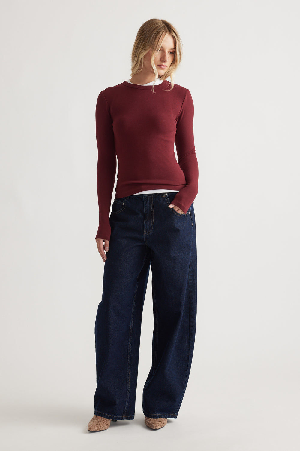 Woman wearing a fitted ruby red long sleeve top layered over white with wide-leg dark blue denim jeans and brown pointed shoes
