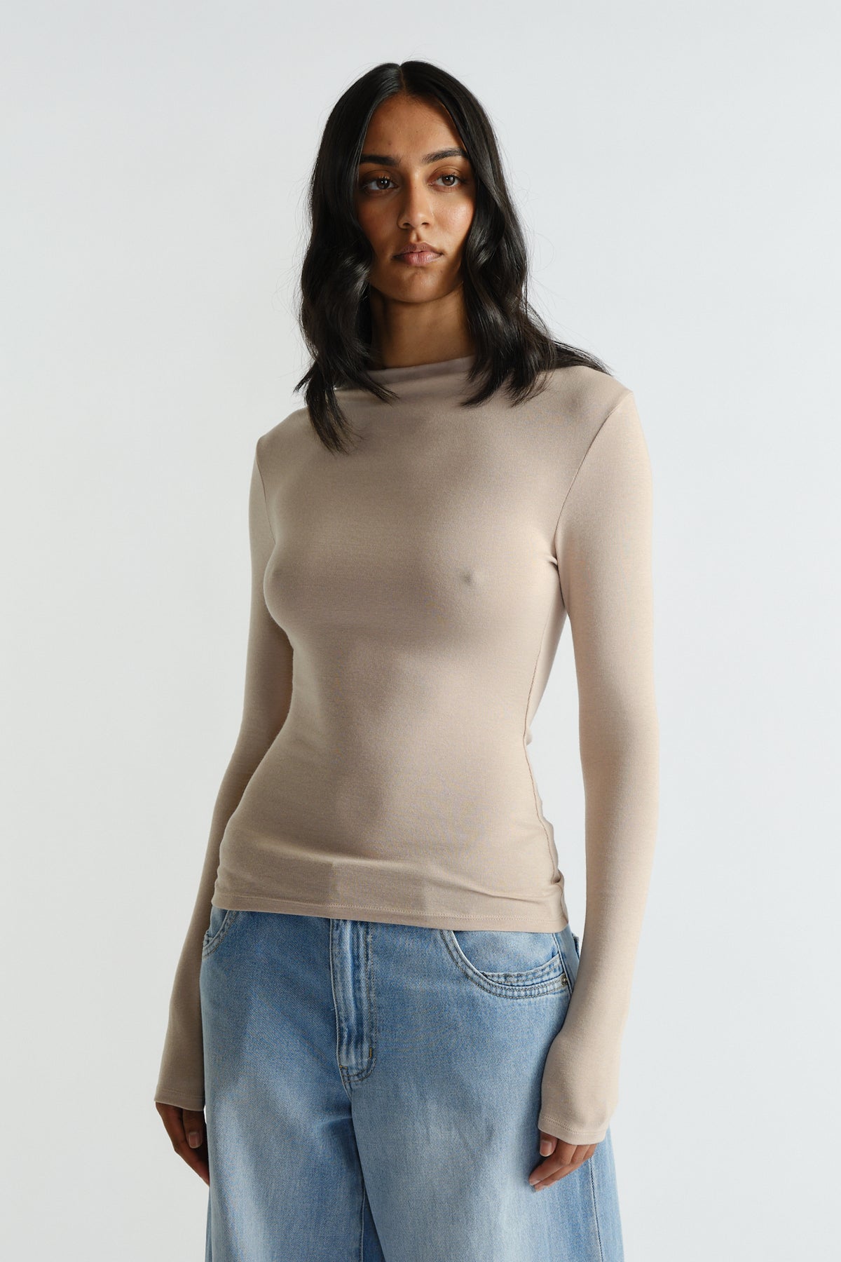 Woman wearing a fitted beige long sleeve top paired with loose light wash denim jeans against a plain background.