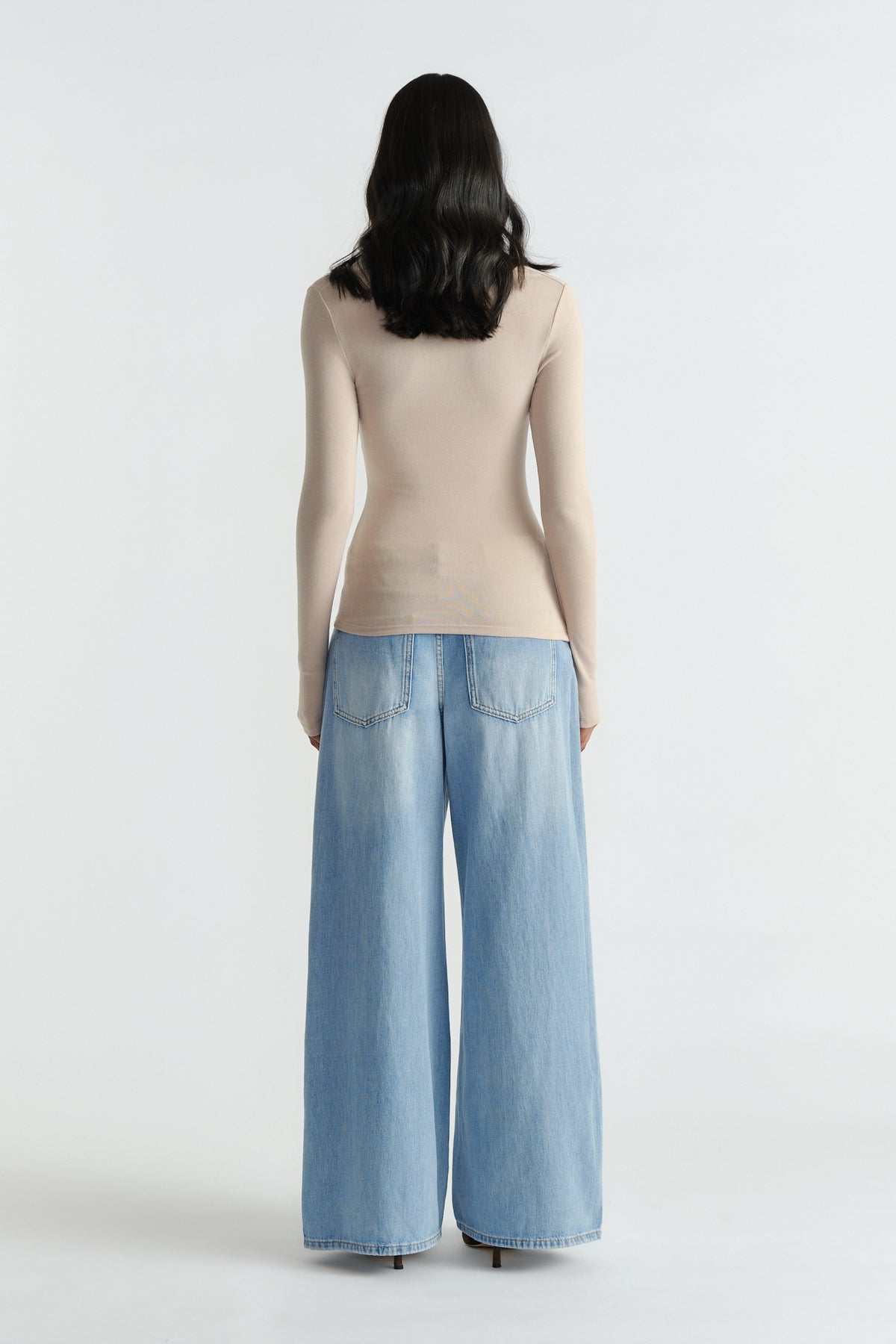 Woman wearing a fitted beige long sleeve top paired with high-rise wide-leg light blue jeans and black heels, back view.