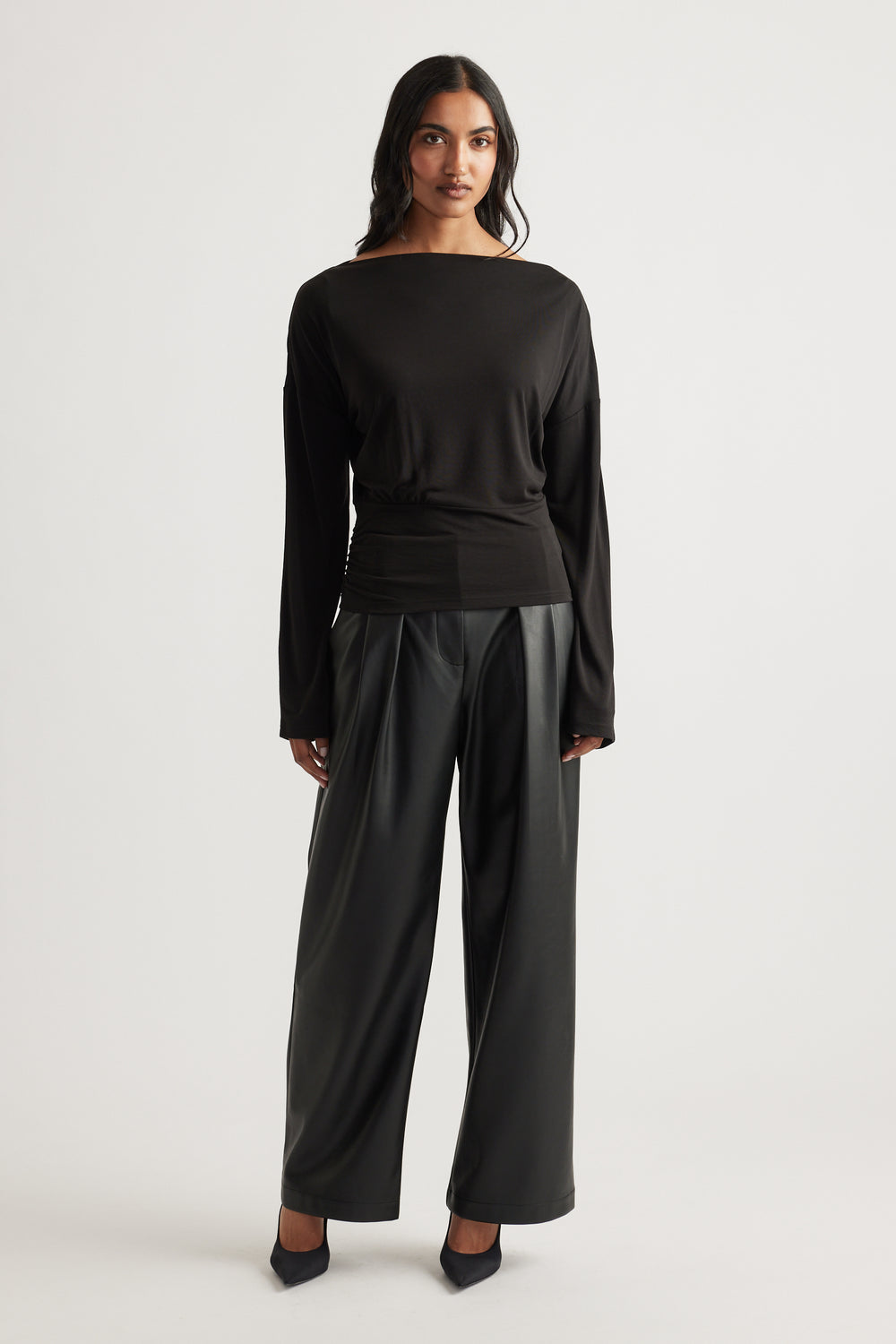 Woman wearing a fitted black long sleeve top with cinched waist paired with wide-leg leather-look pants and heels