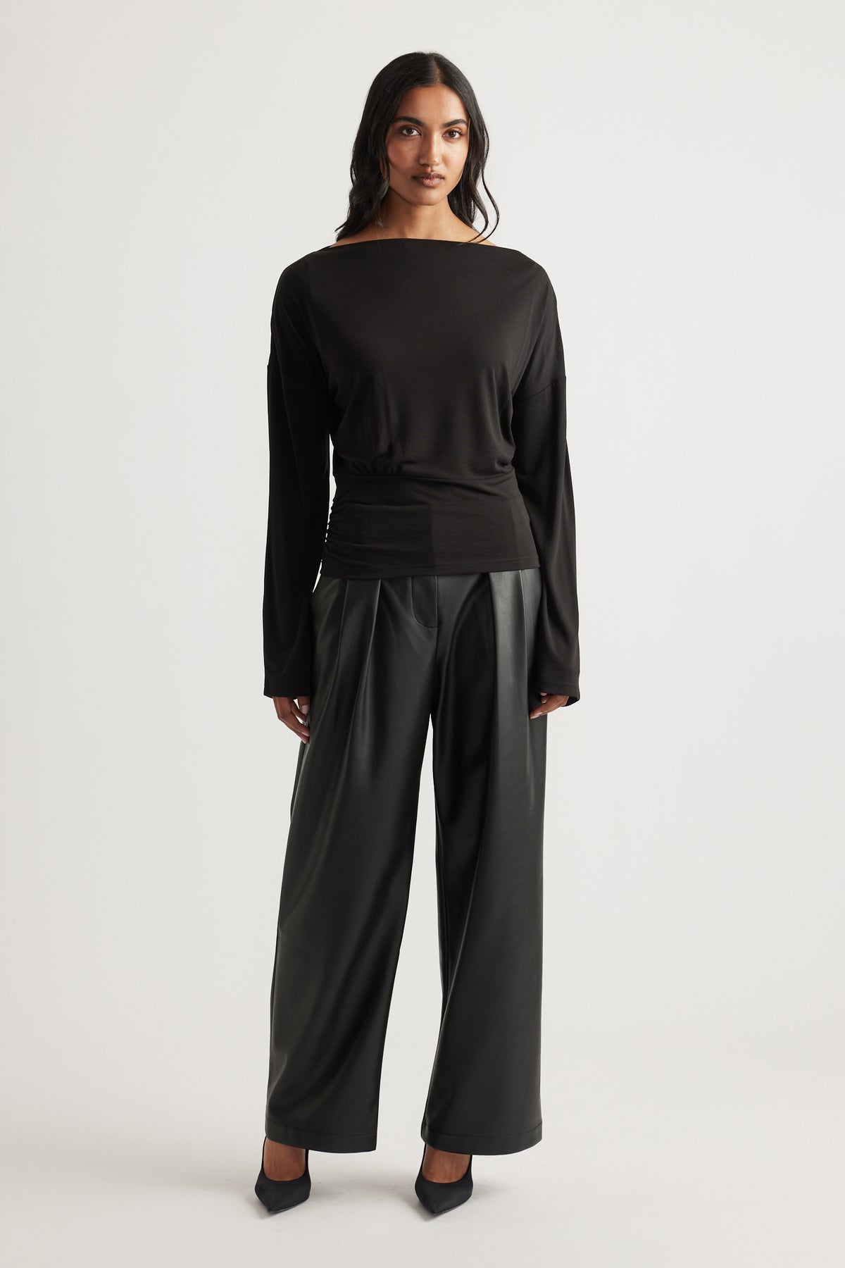 Woman wearing a fitted black long sleeve top with cinched waist paired with wide-leg leather-look pants and heels