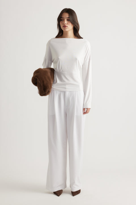 Woman wearing a fitted white long sleeve top with wide-leg white pants, holding a brown textured jacket for contrast