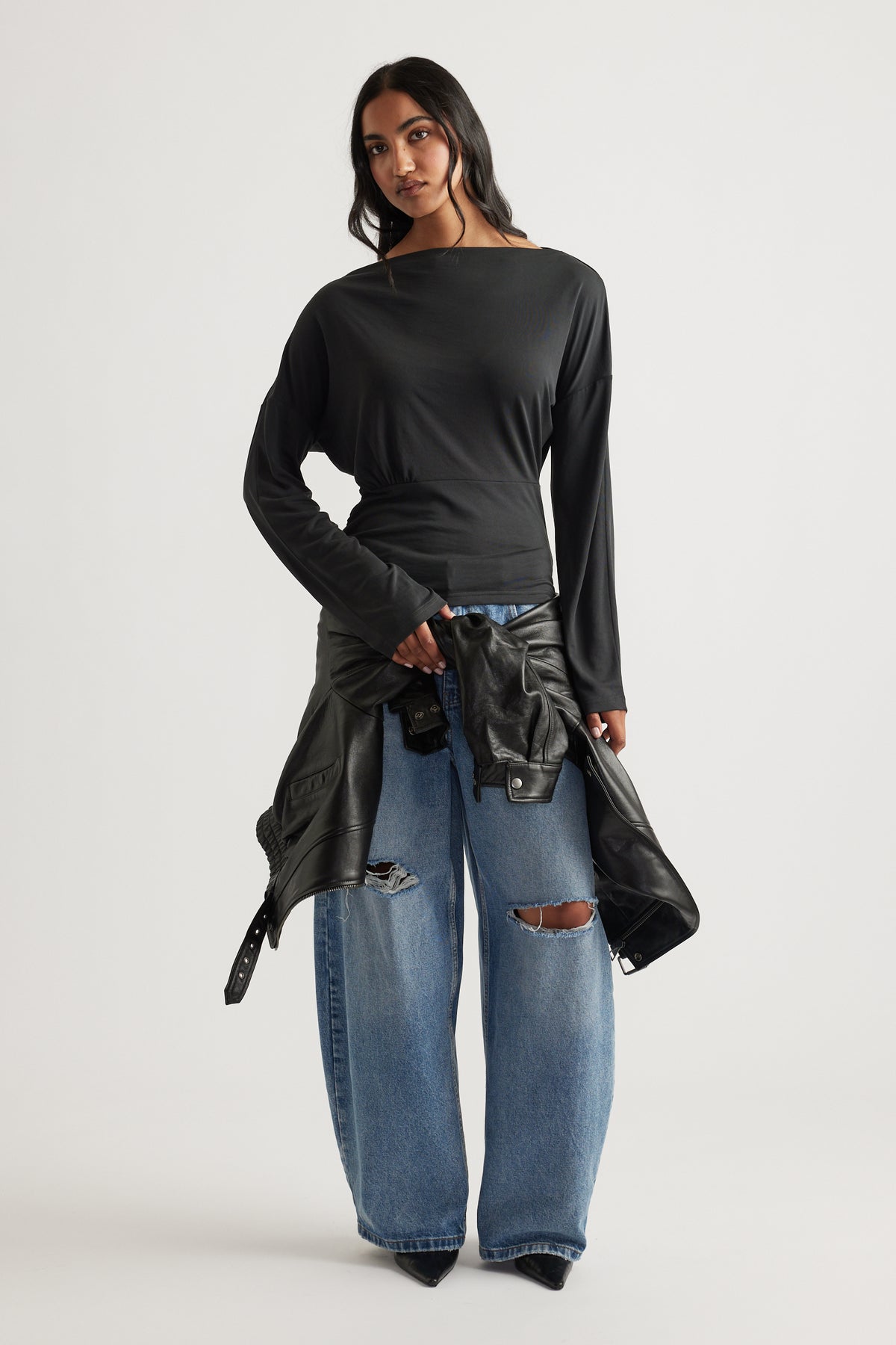 Woman wearing a fitted black long sleeve top paired with wide-leg distressed blue jeans and holding a leather jacket