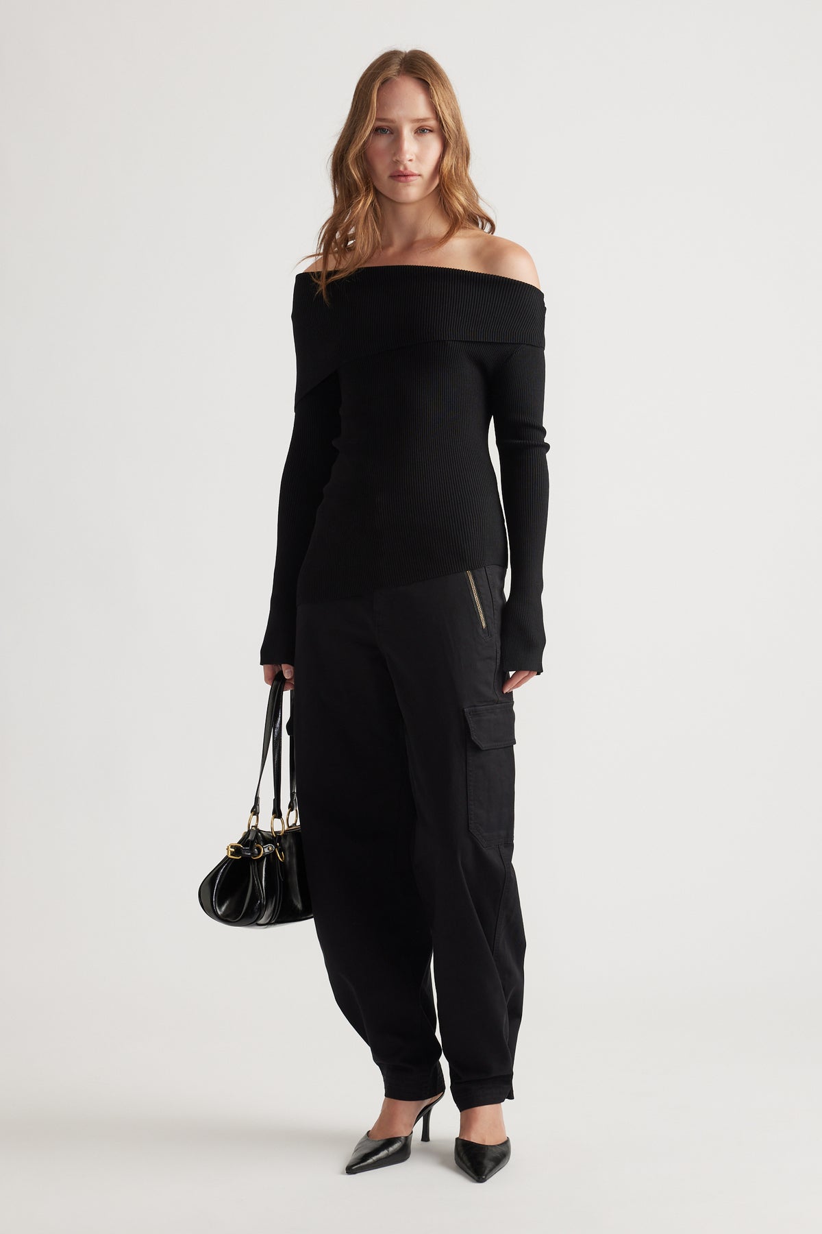 Woman wearing a sleek black off-shoulder knit top paired with relaxed cargo pants and pointed heels, holding a leather bag.
