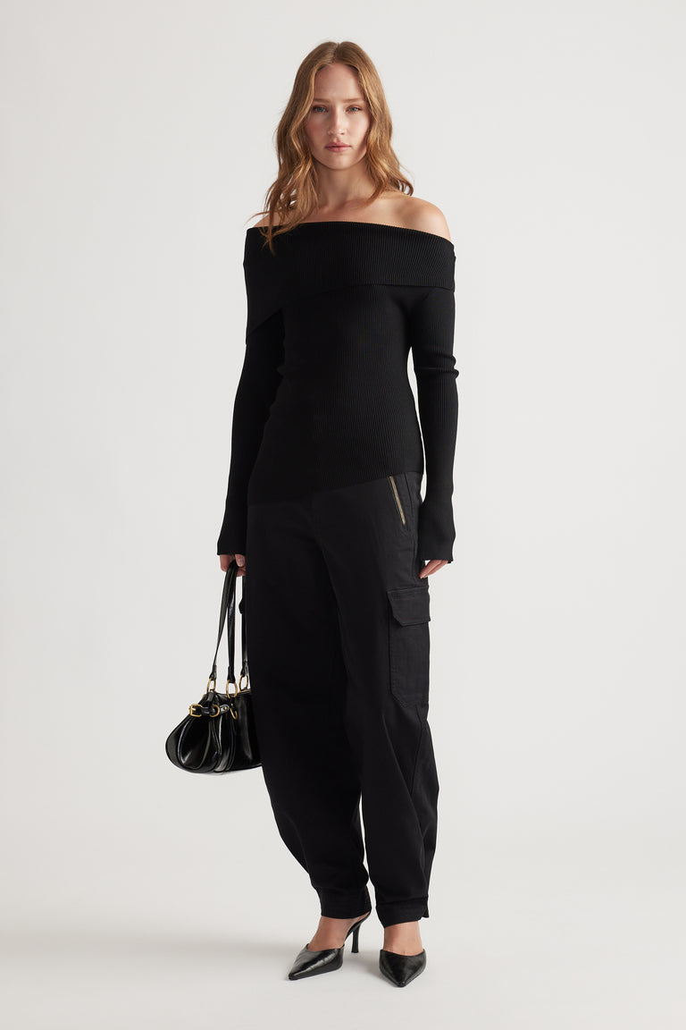 Woman wearing a sleek black off-shoulder knit top paired with relaxed cargo pants and pointed heels, holding a leather bag.