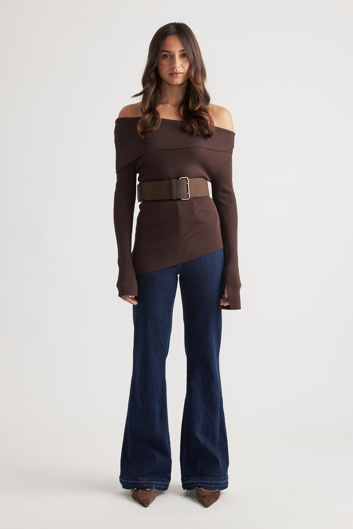 Woman wearing a rich mocha off-shoulder knit top with a wide belt, paired with dark flared denim jeans and pointed shoes