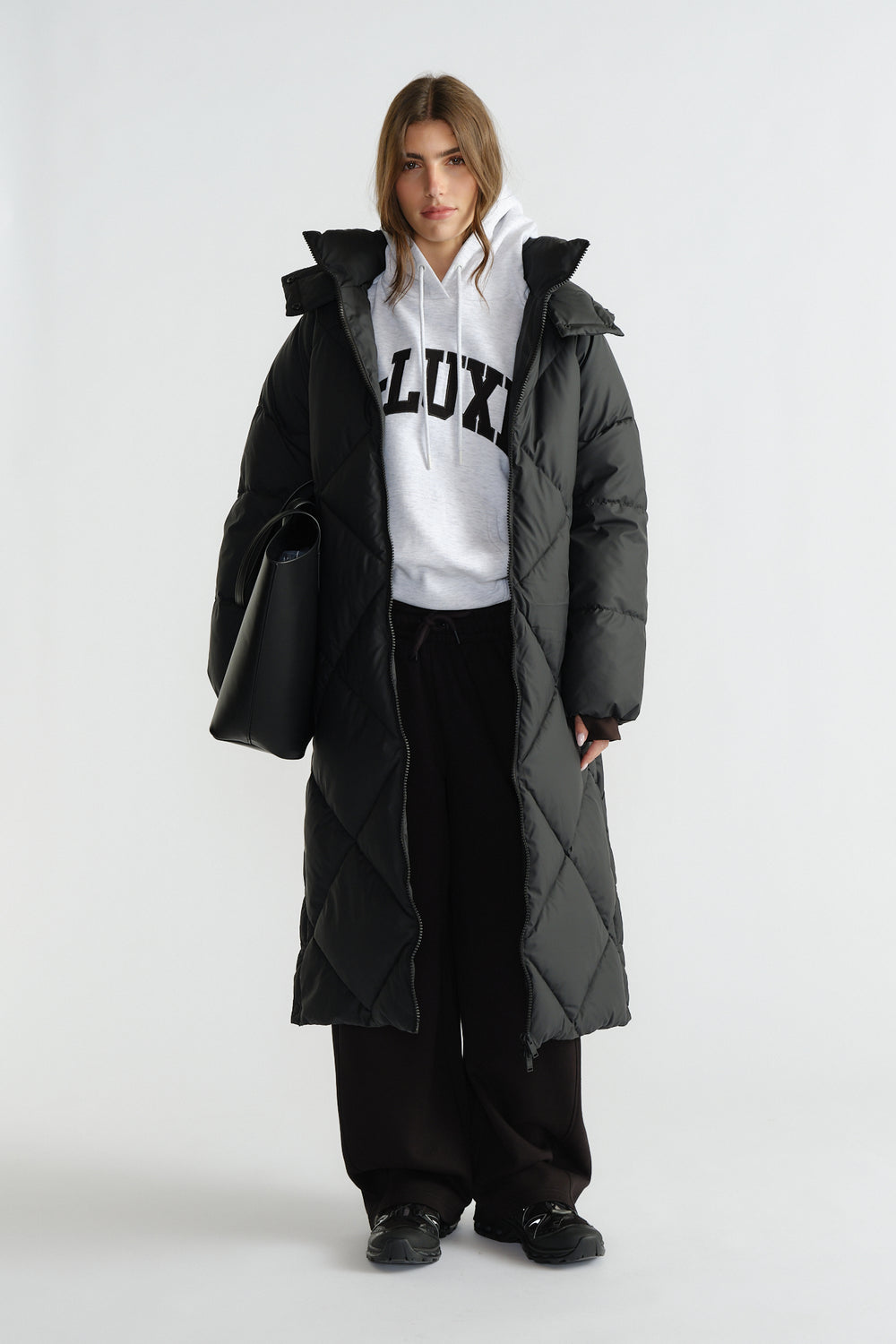 Woman wearing a long black quilted puffer coat over a white hoodie and wide-leg black pants, carrying a large black tote bag.
