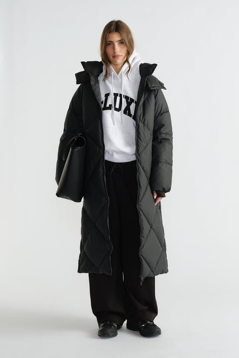 Woman wearing a long black quilted puffer coat over a white hoodie and wide-leg black pants, carrying a large black tote bag.