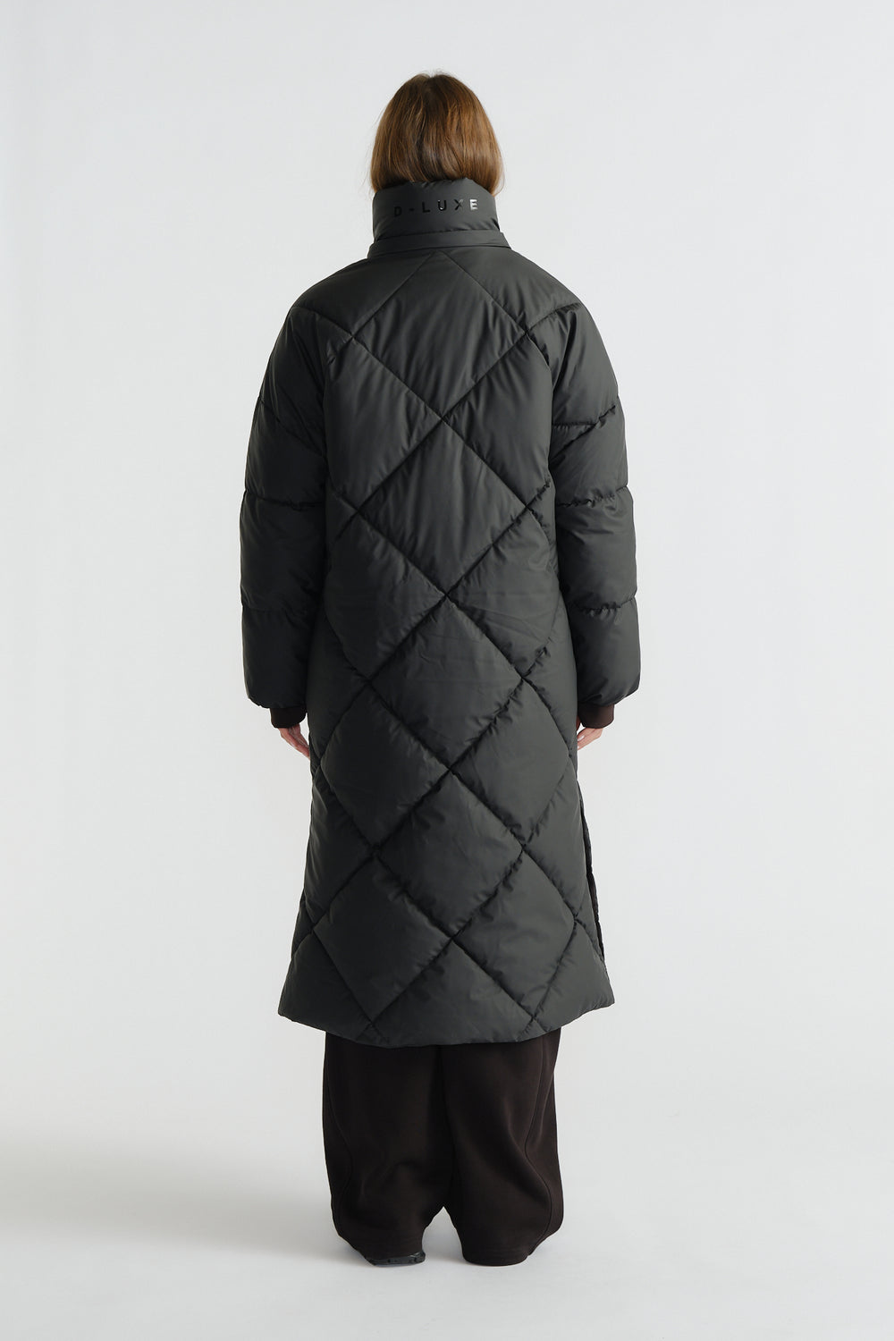 Woman seen from behind wearing a long black quilted puffer coat with diamond stitching and a high collar, paired with wide pants