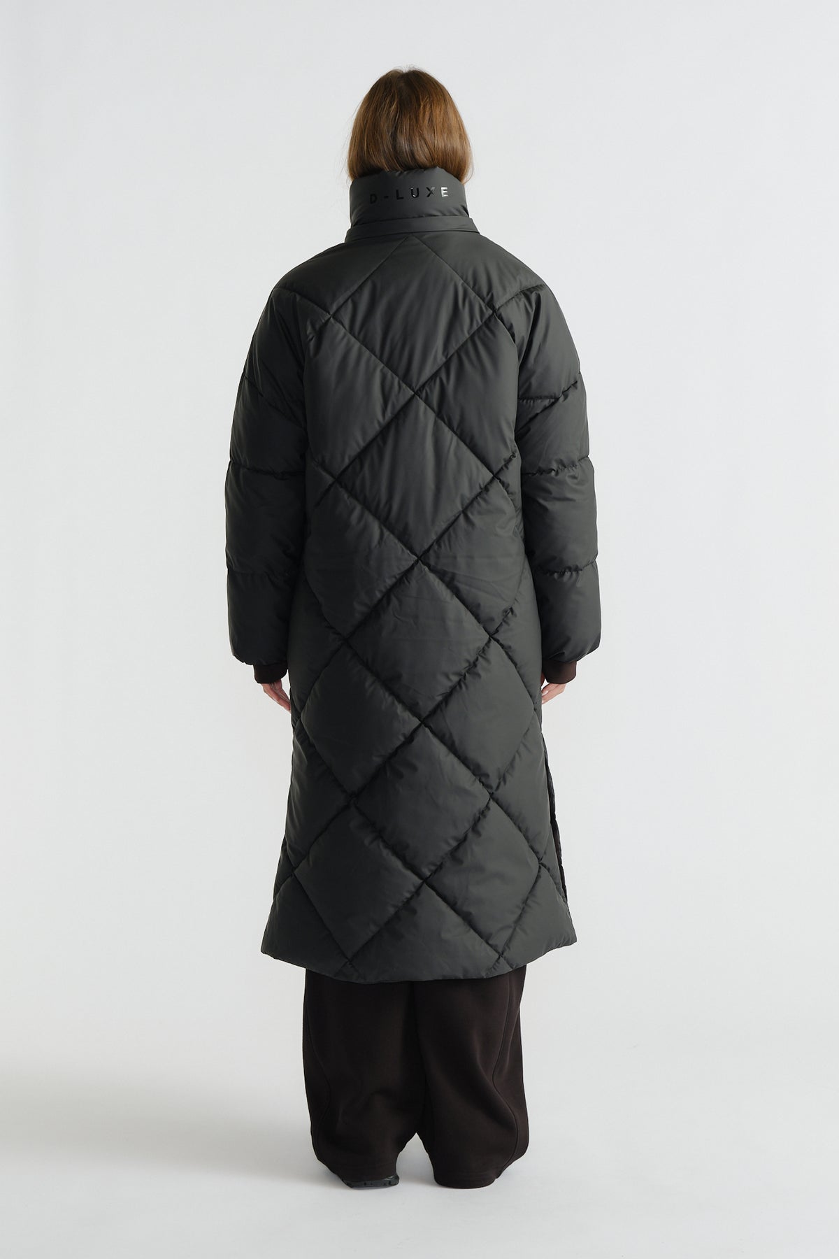 Woman seen from behind wearing a long black quilted puffer coat with diamond stitching and a high collar, paired with wide pants