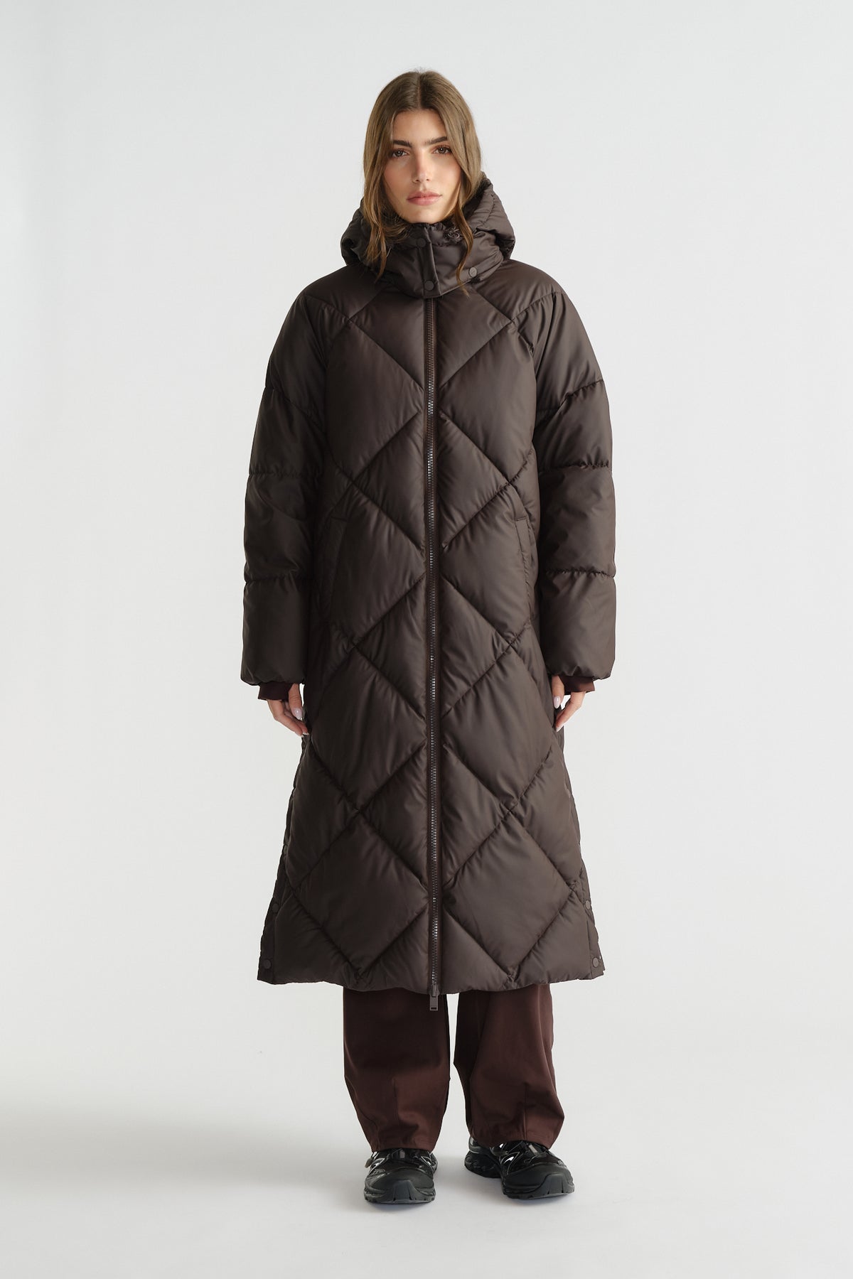 Woman wearing a long, brown quilted puffer coat with a hood, styled with loose brown pants and black shoes