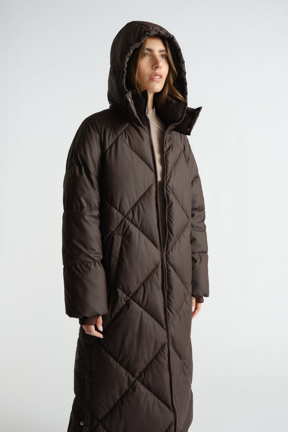 Woman wearing a long chocolate brown quilted puffer coat with hood and side pockets, styled with a beige knit top