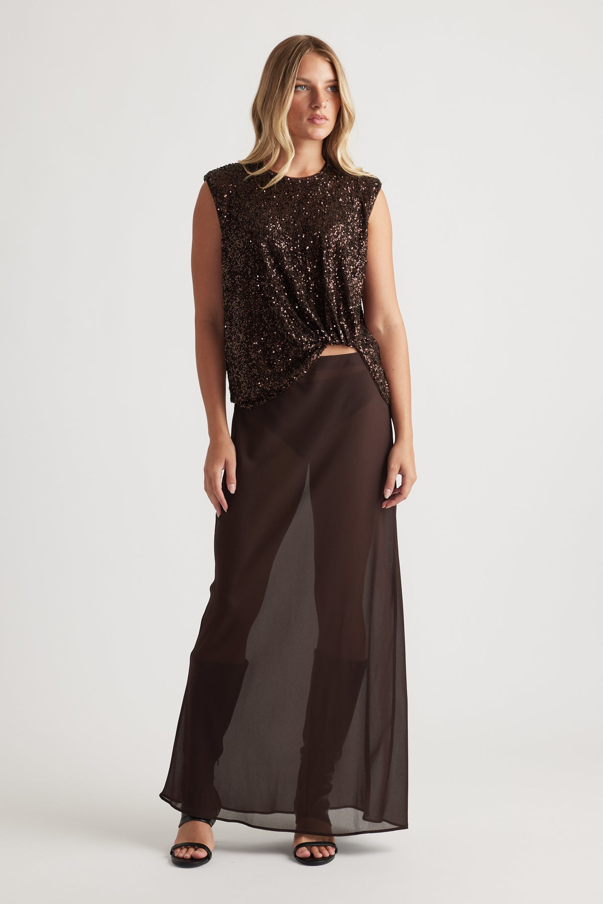 Woman wearing a sleeveless dark brown sequined top paired with sheer wide-leg pants and black sandals on a plain background