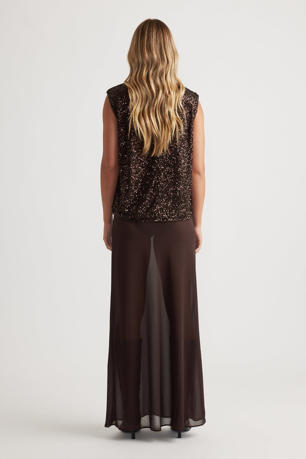 Woman with long wavy hair wearing a sleeveless dark sequined top and flowing sheer brown wide-leg pants