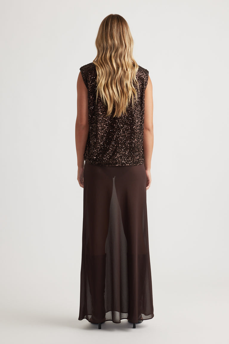 Woman with long wavy hair wearing a sleeveless dark sequined top and flowing sheer brown wide-leg pants