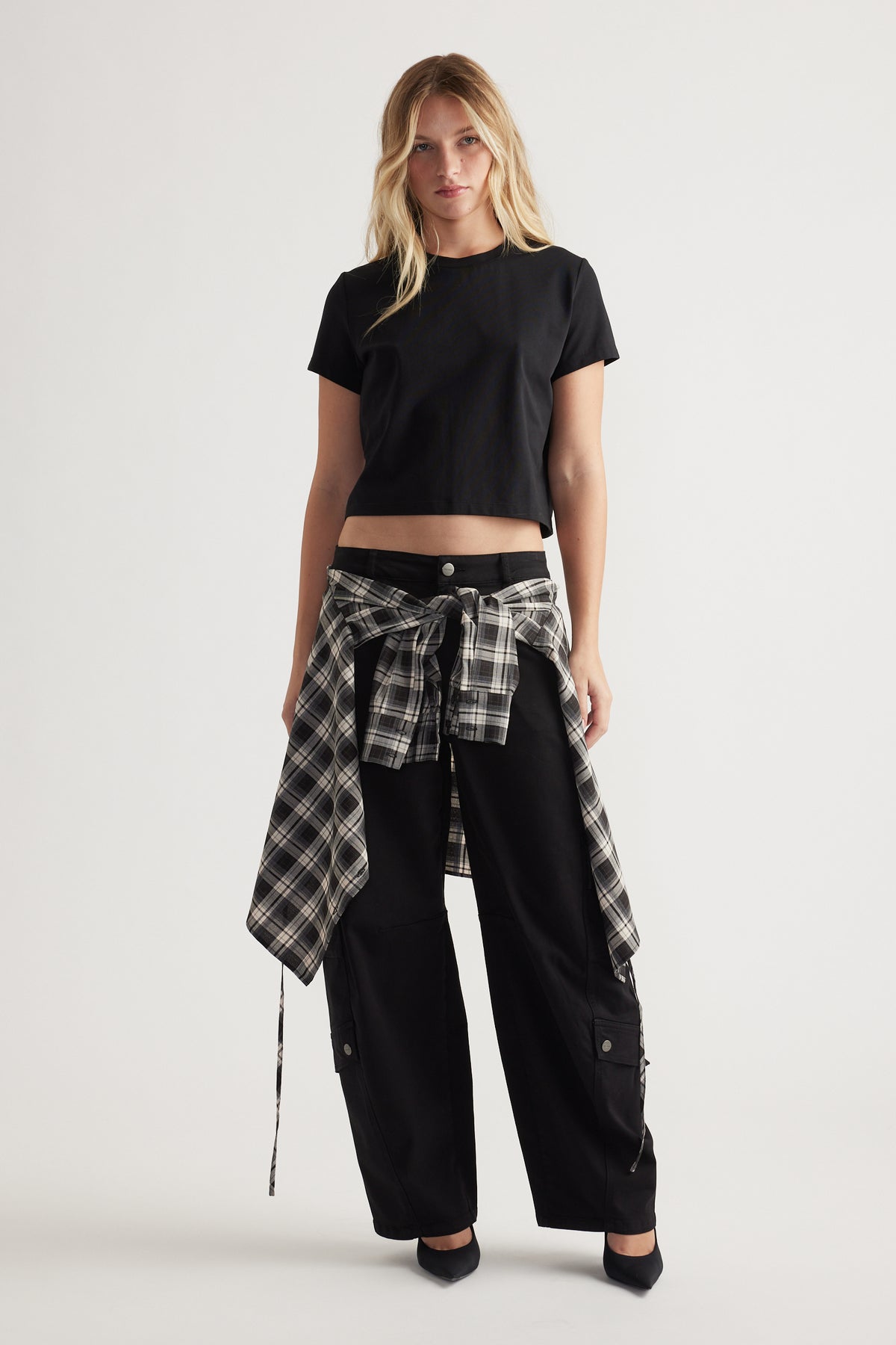 Woman wearing a black cropped tee with relaxed black trousers and a black-and-white checked shirt tied at the waist.