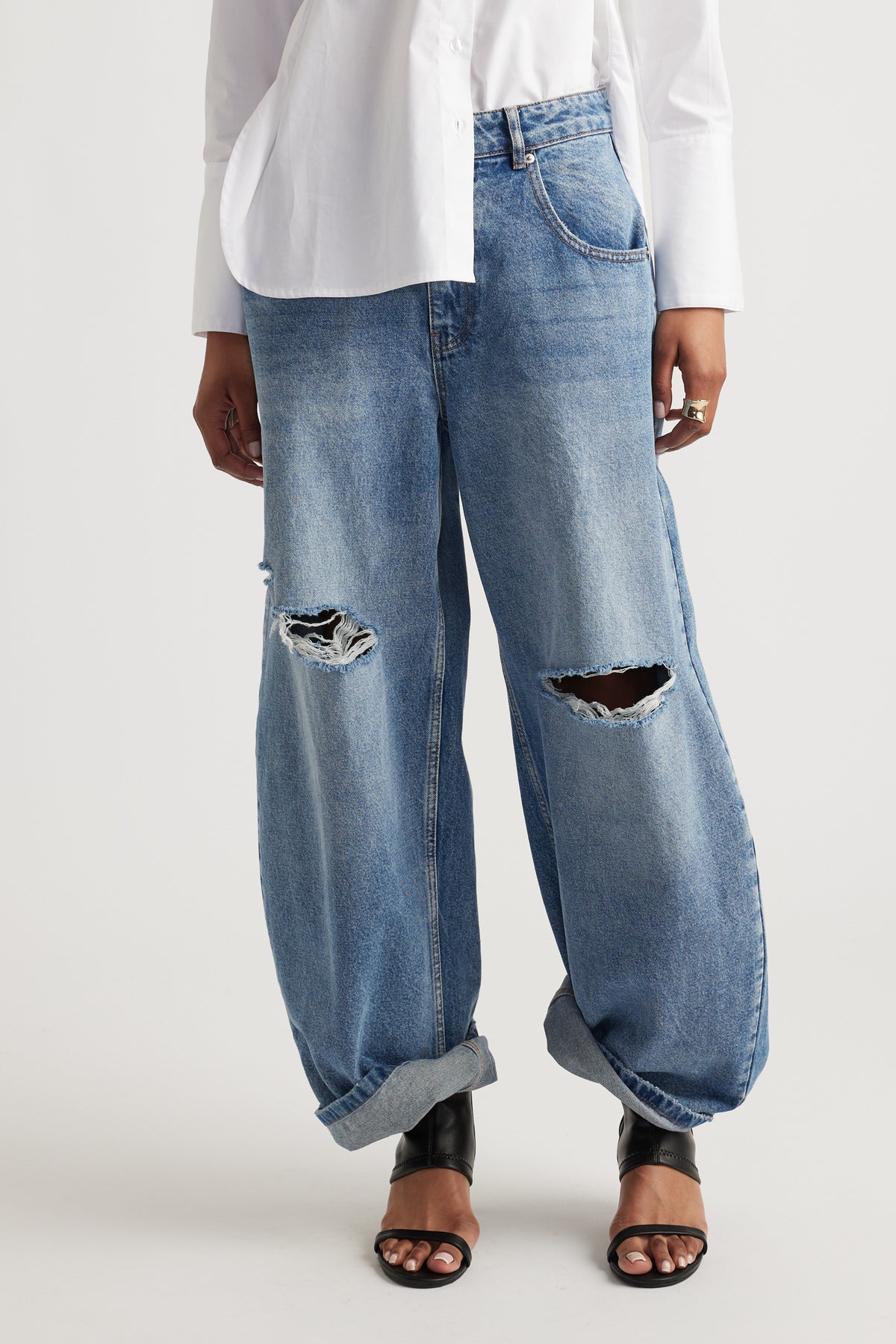 Woman wearing light blue high rise horseshoe jeans with distressed knees, a white shirt and black sandals