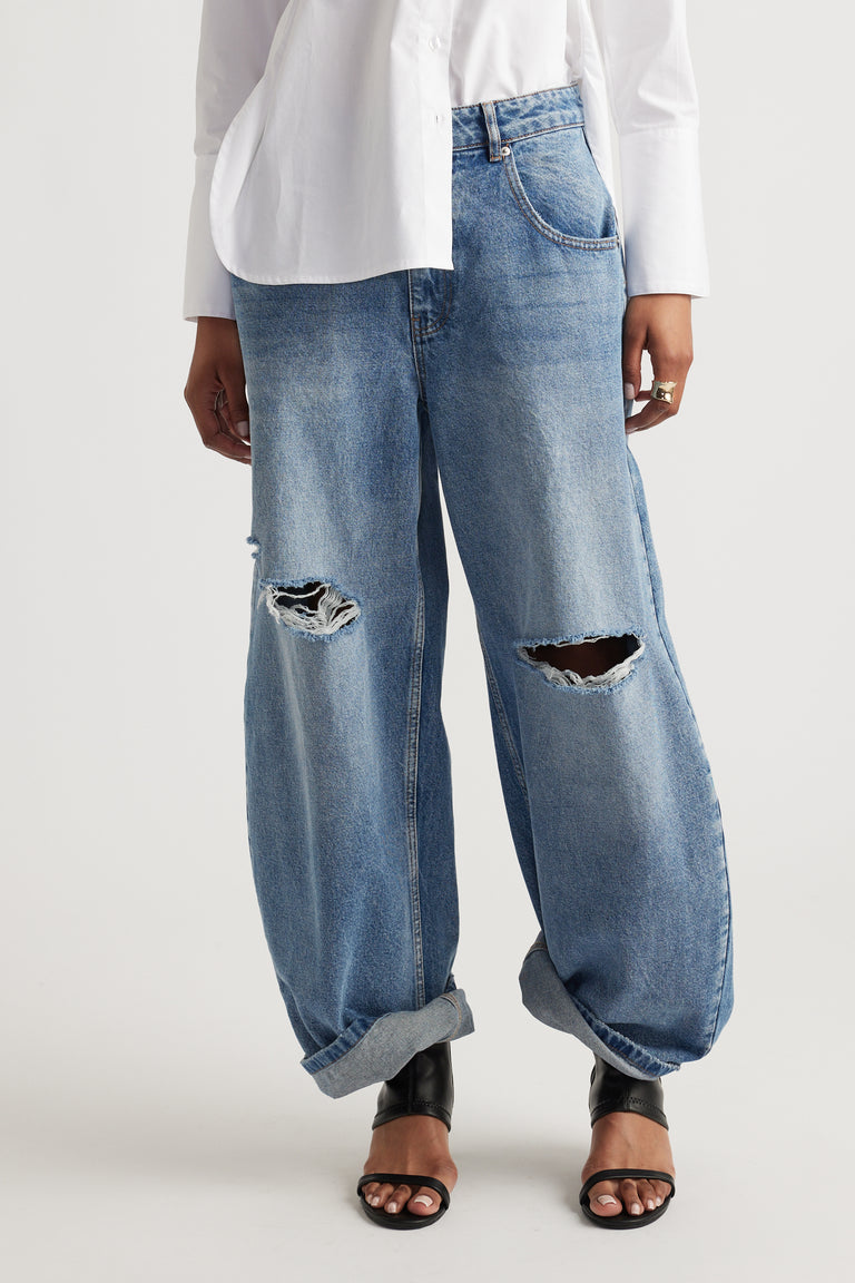 Woman wearing light blue high rise horseshoe jeans with distressed knees, a white shirt and black sandals