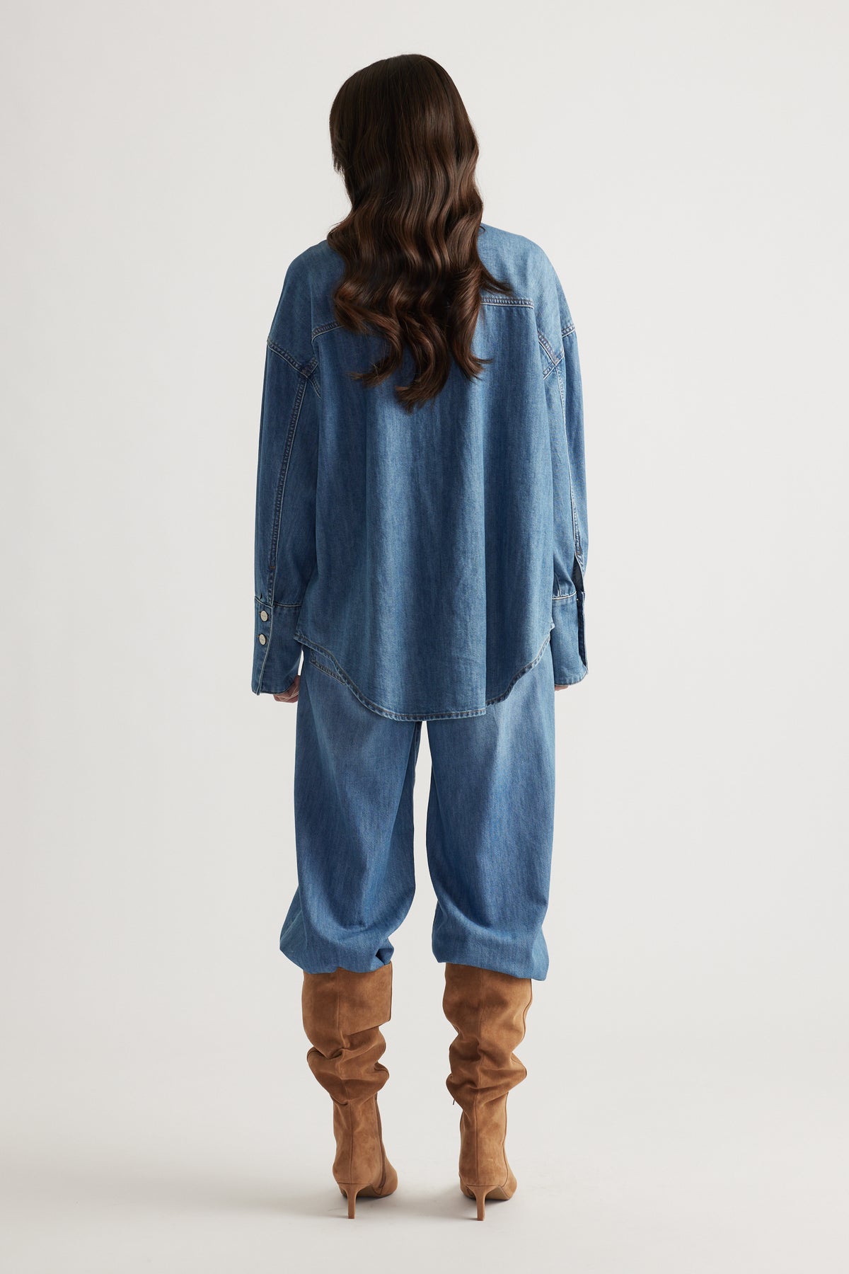 Woman wearing relaxed fit blue denim shirt and loose denim pants paired with slouchy tan suede boots, back view.