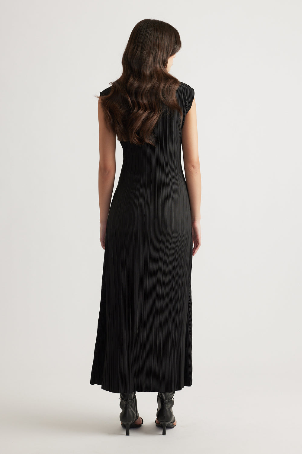 Woman wearing a sleek black pleated maxi dress with cap sleeves and a flowing silhouette paired with black heeled boots.