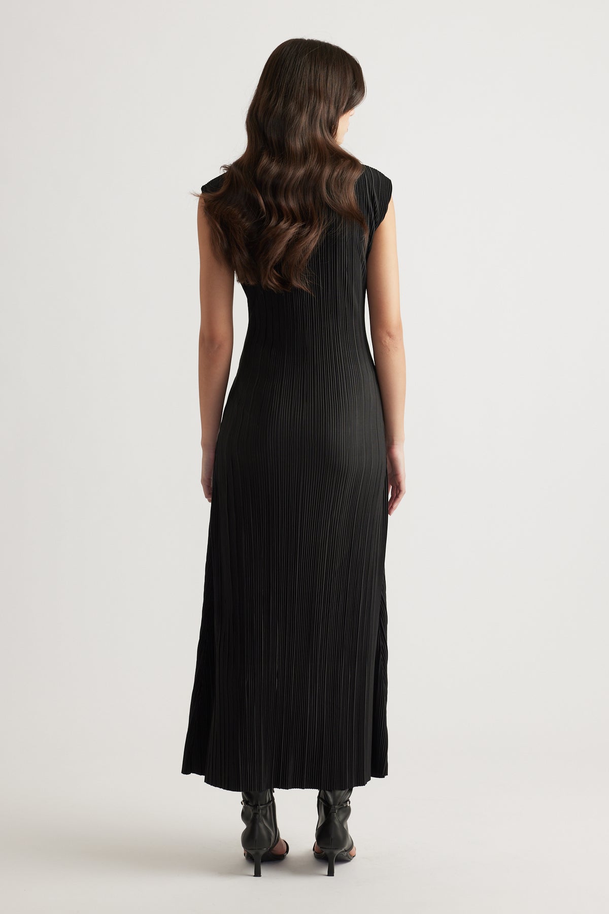 Woman wearing a sleek black pleated maxi dress with cap sleeves and a flowing silhouette paired with black heeled boots.