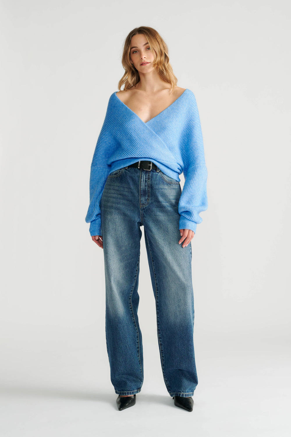 Woman wearing a relaxed blue crossover knit jumper paired with high-rise wide-leg denim jeans and black pointed shoes.