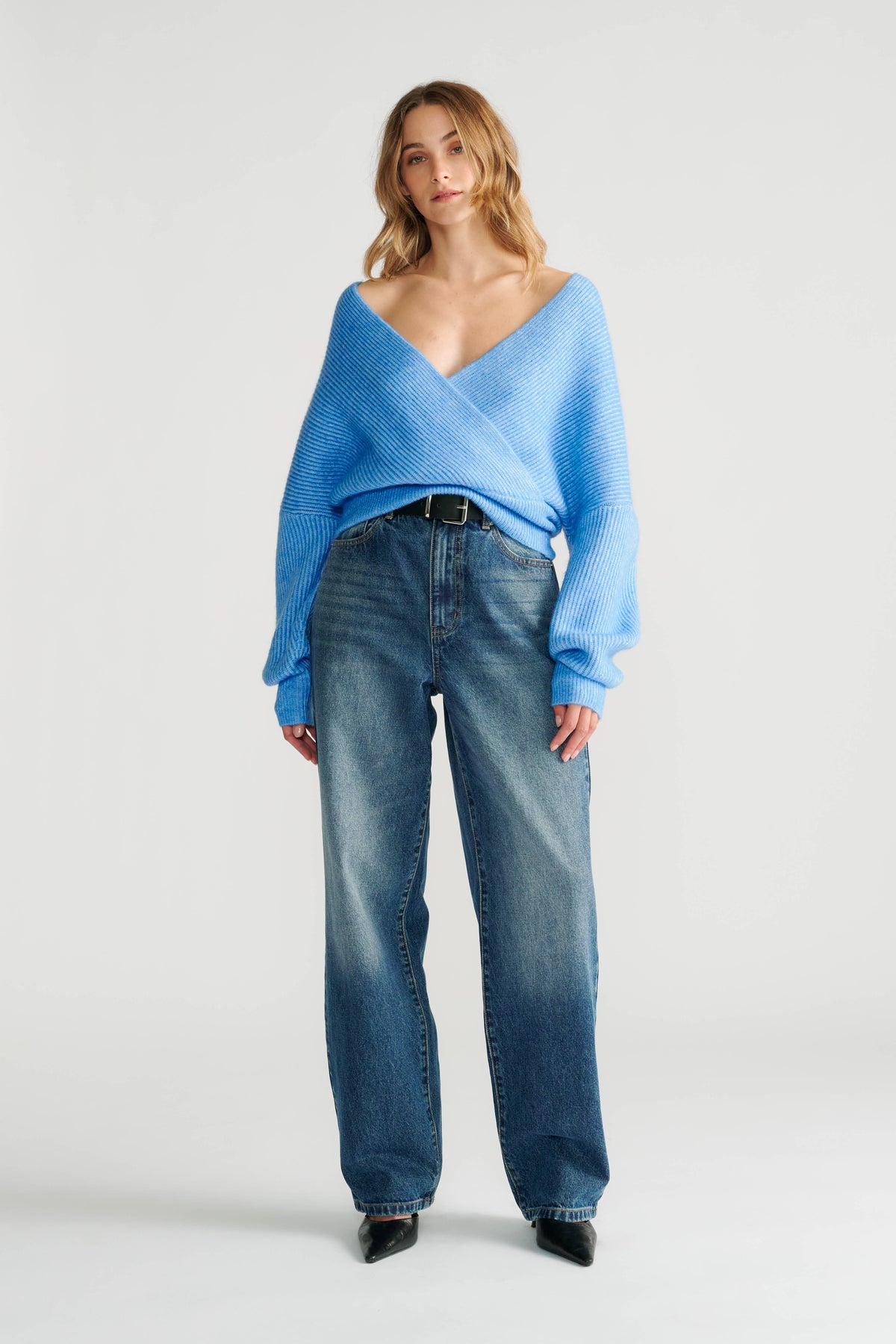 Woman wearing a relaxed blue crossover knit jumper paired with high-rise wide-leg denim jeans and black pointed shoes.