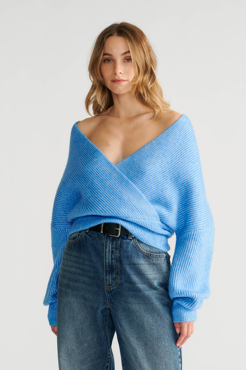 Woman wearing a soft blue crossover knit jumper with voluminous sleeves paired with high-waisted blue jeans and a black belt.