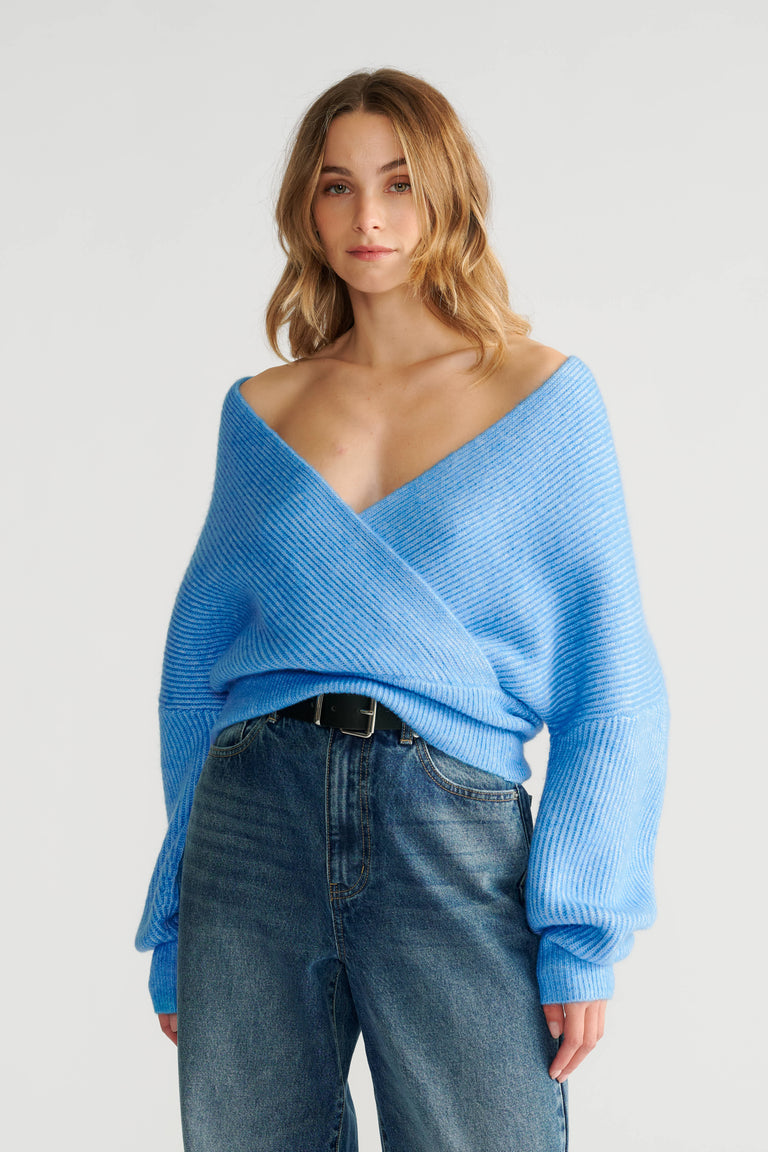 Woman wearing a soft blue crossover knit jumper with voluminous sleeves paired with high-waisted blue jeans and a black belt.