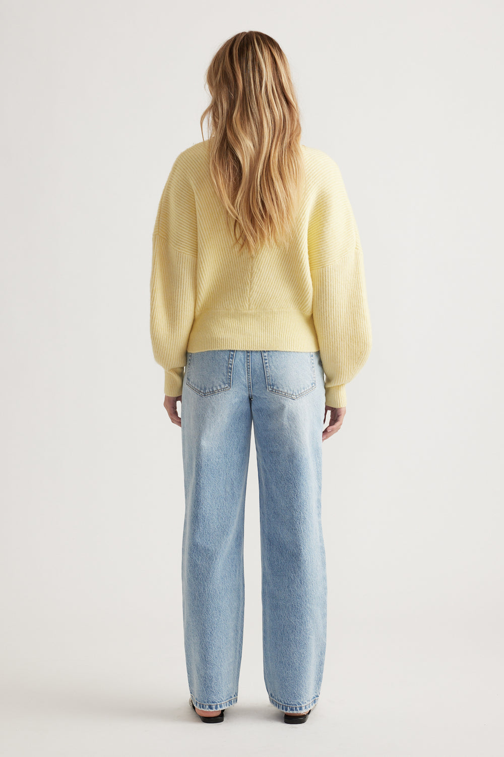 Woman seen from behind wearing a soft yellow crossover knit jumper paired with relaxed light-wash blue jeans