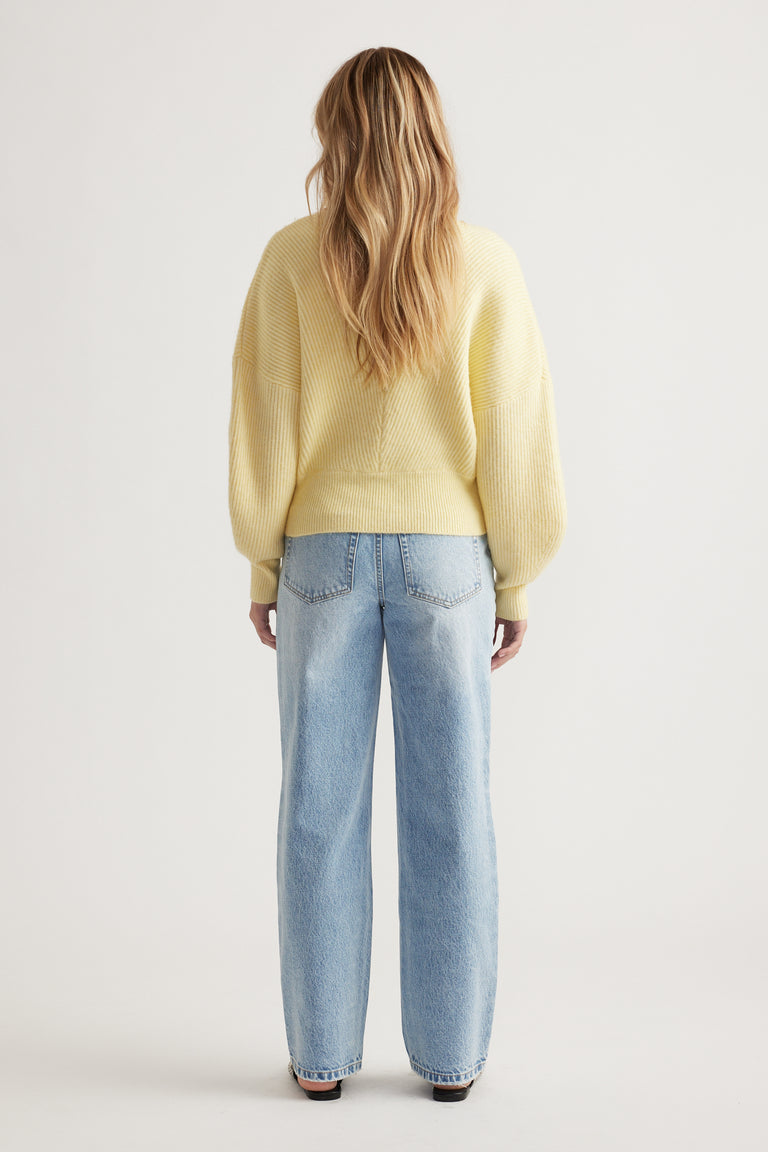 Woman seen from behind wearing a soft yellow crossover knit jumper paired with relaxed light-wash blue jeans