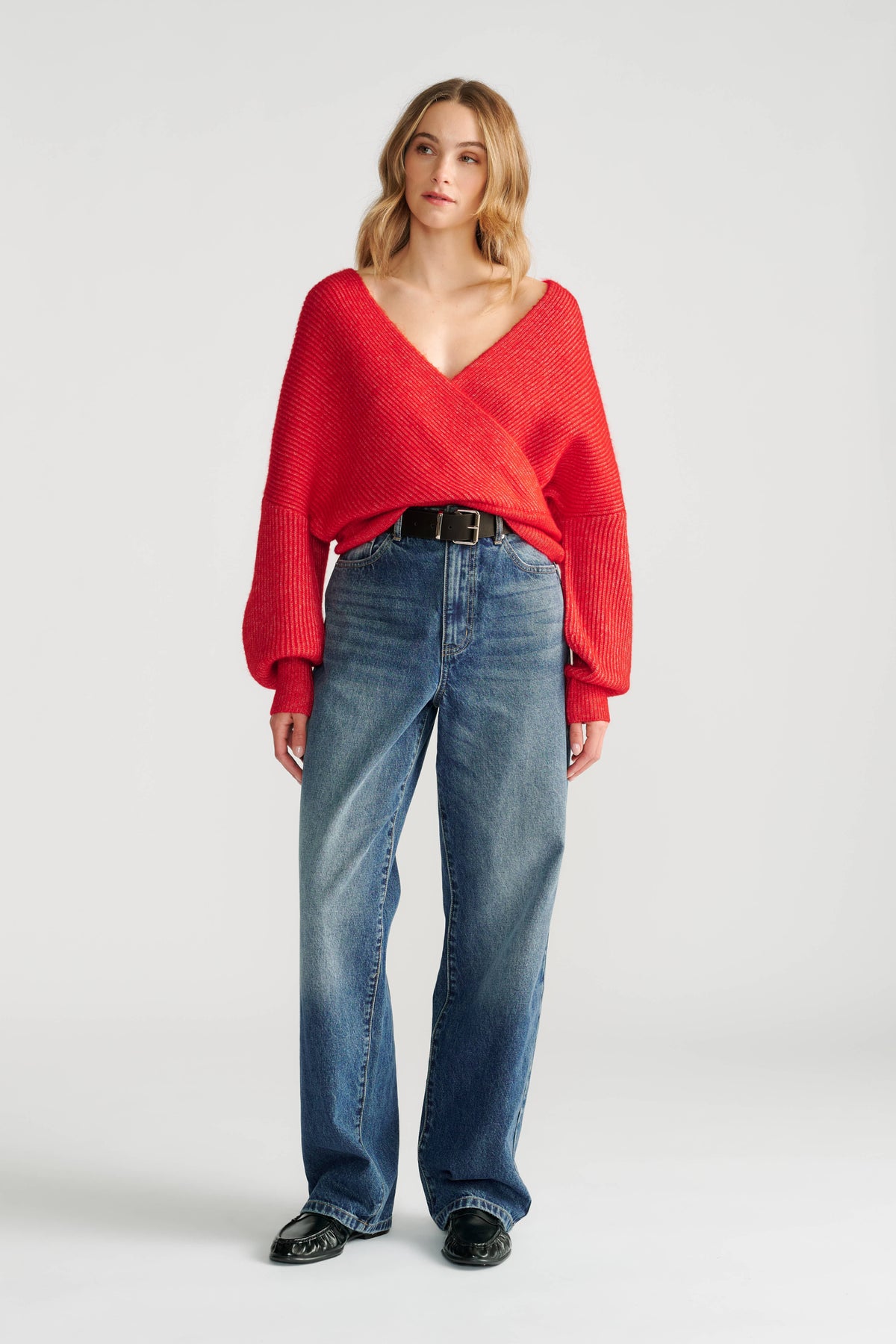 Woman wearing a bright red crossover knit jumper styled with high-rise relaxed fit blue jeans and black belt and shoes.