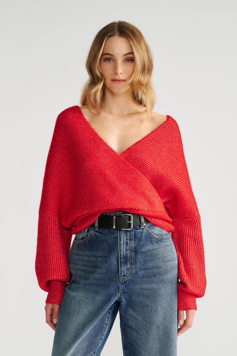 Woman wearing a vibrant red crossover knit jumper with voluminous sleeves paired with high-waisted blue jeans and a black belt.