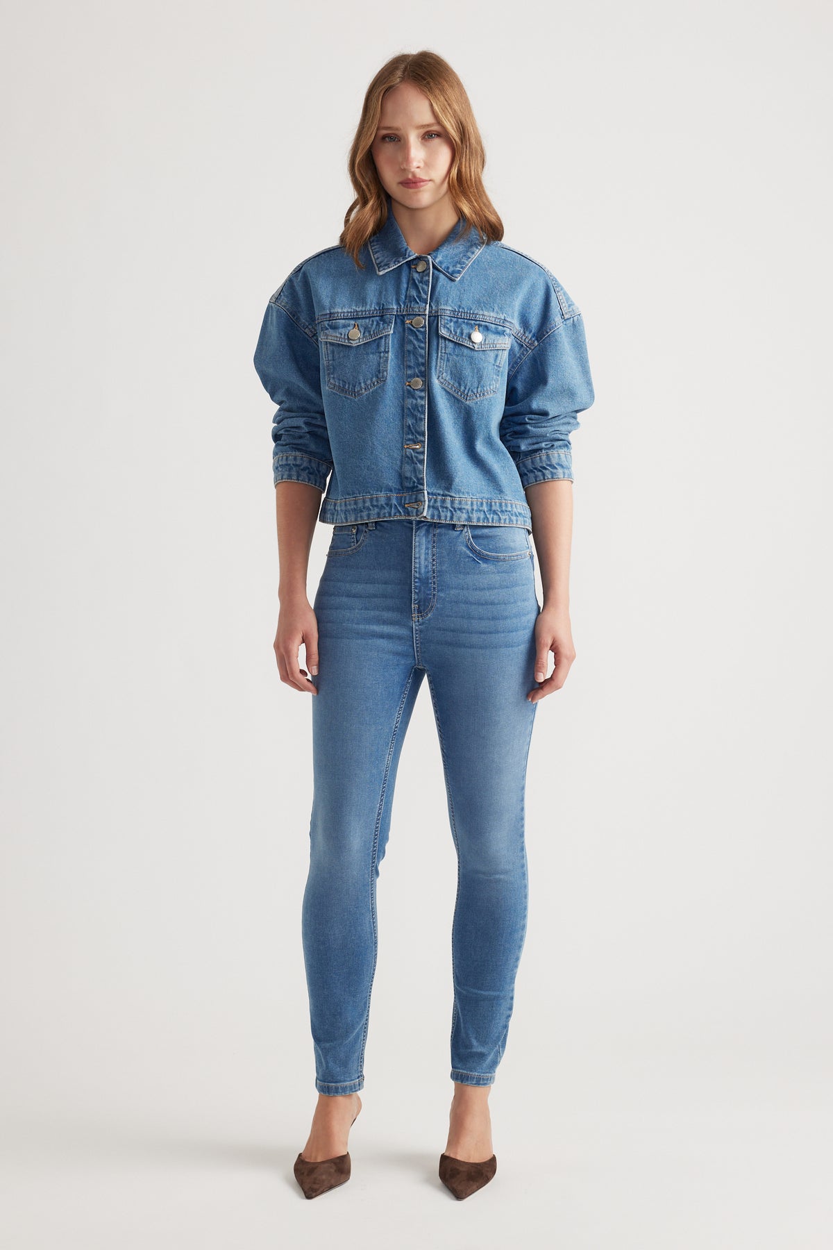 Woman wearing a mid-blue cropped denim trucker jacket paired with high-waisted skinny jeans and brown pointed heels against white background