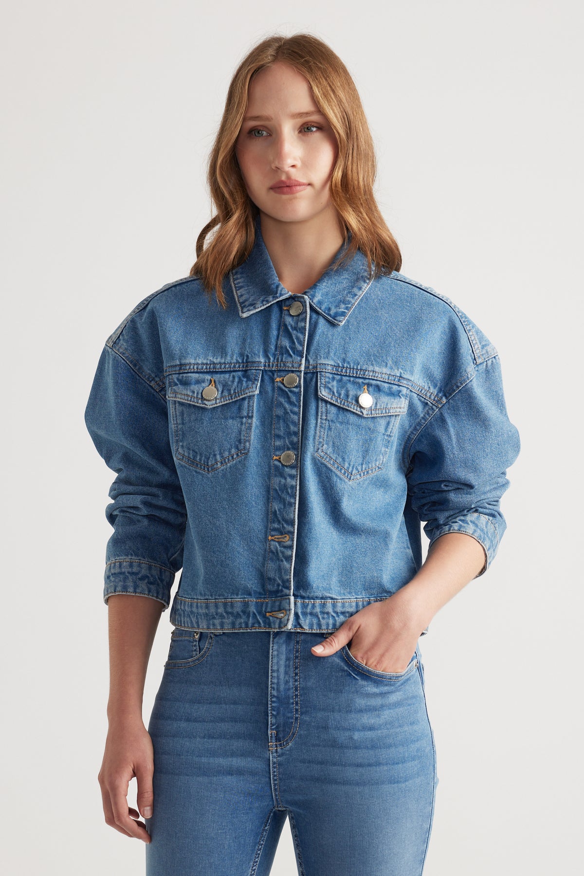 Woman wearing a mid-blue cropped denim jacket with button details paired with high-rise skinny jeans against white background