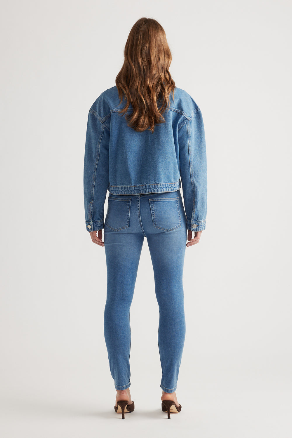 Woman shown from behind wearing mid-blue cropped denim jacket paired with matching high-waisted skinny jeans and brown heels