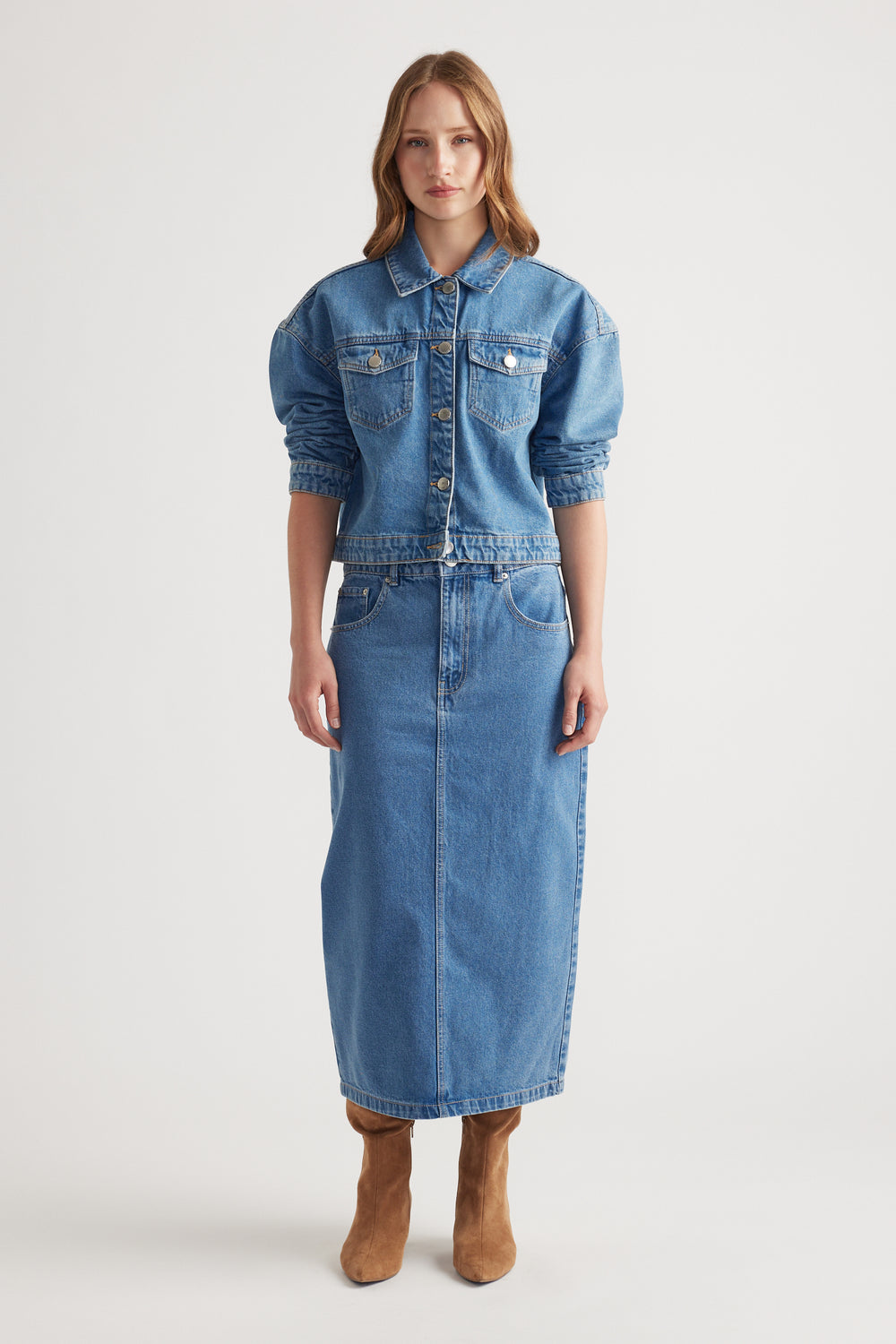 Woman wearing mid-blue denim midi skirt with front pockets paired with matching cropped denim jacket and tan boots