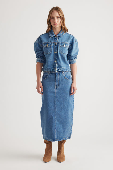 Woman wearing mid-blue denim midi skirt with front pockets paired with matching cropped denim jacket and tan boots