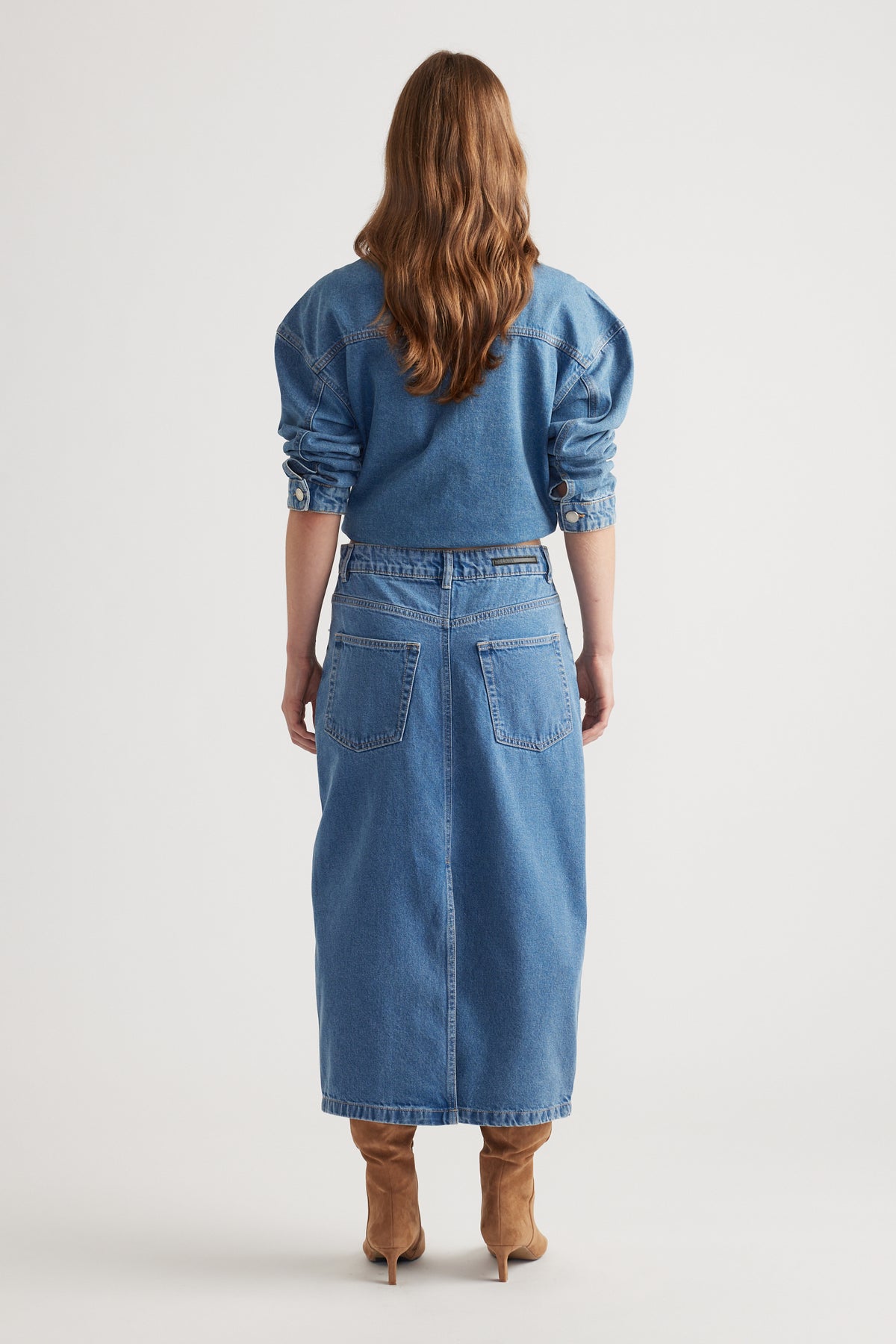 Woman wearing a mid-blue denim midi skirt with back pockets, paired with a matching cropped denim jacket and tan boots