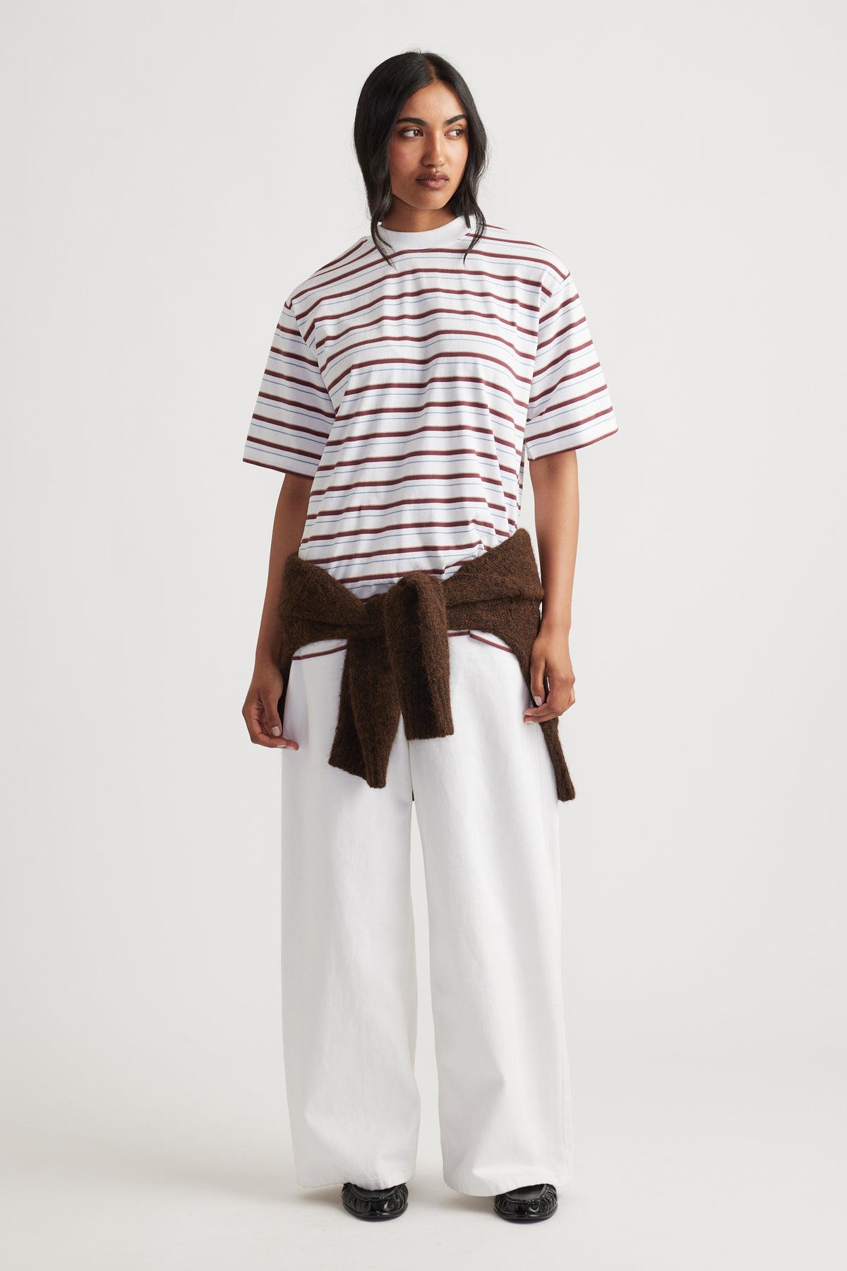Woman wearing Isabella Oversized Stripe Tee in white with brown stripes, styled with wide-leg white pants and a brown sweater by DECJUBA