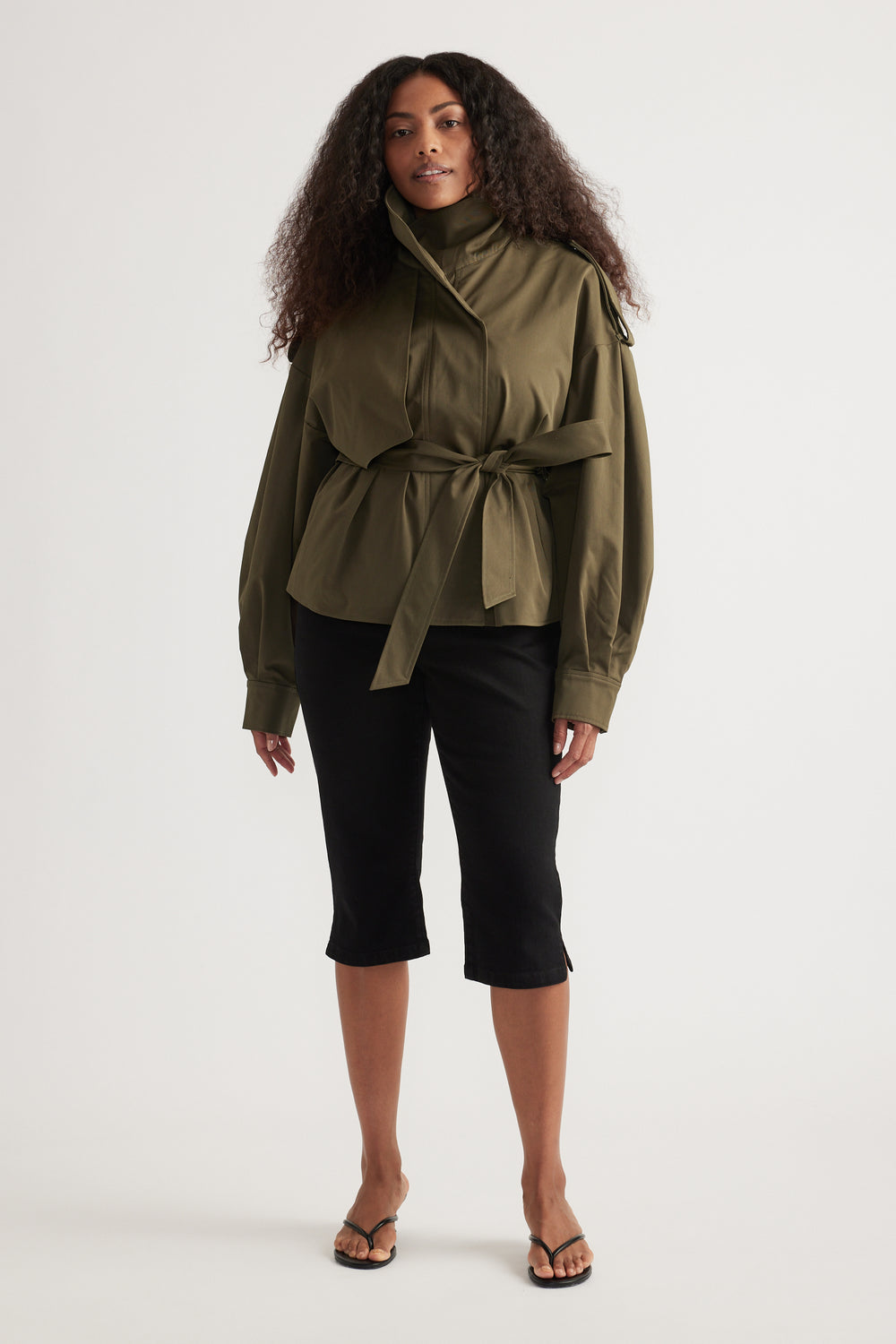 Woman wearing an olive green cropped trench coat with a tie waist, black cropped pants and thong sandals