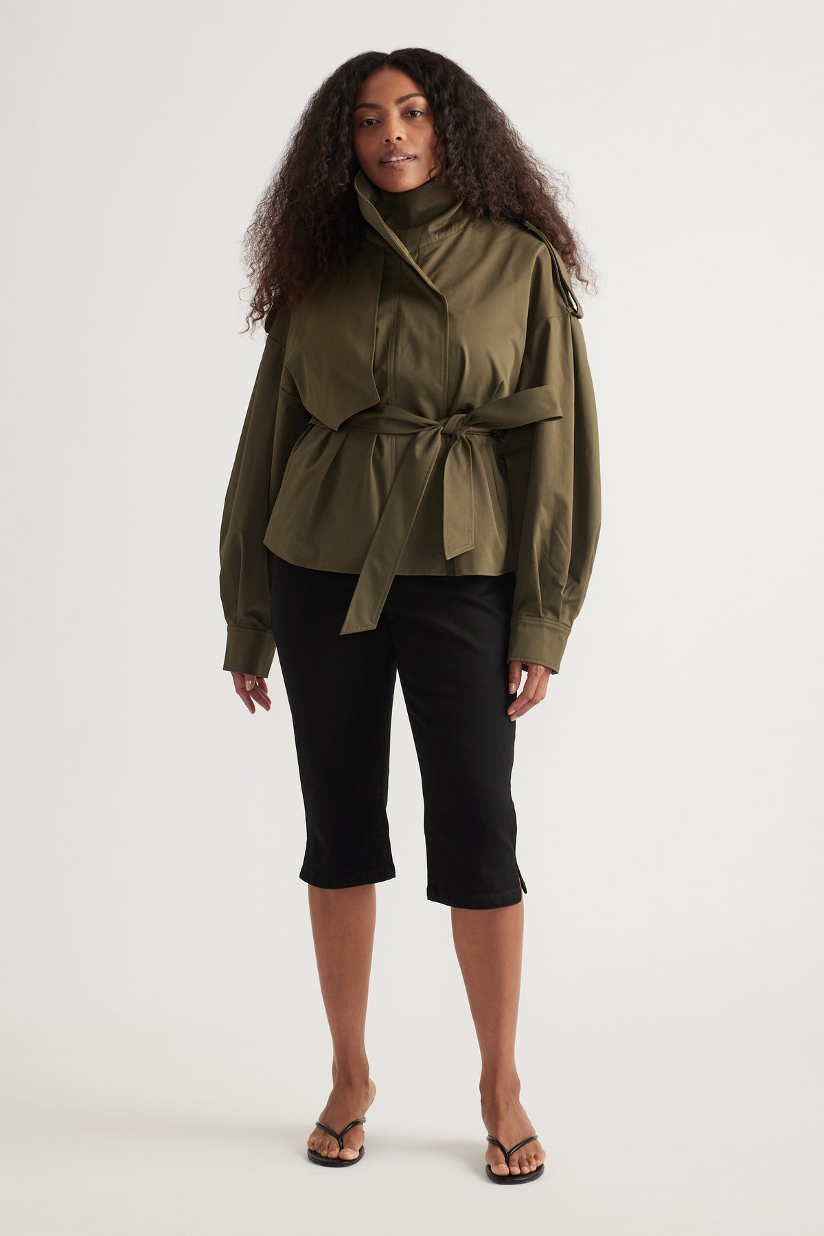 Woman wearing an olive green cropped trench coat with a tie waist, black cropped pants and thong sandals