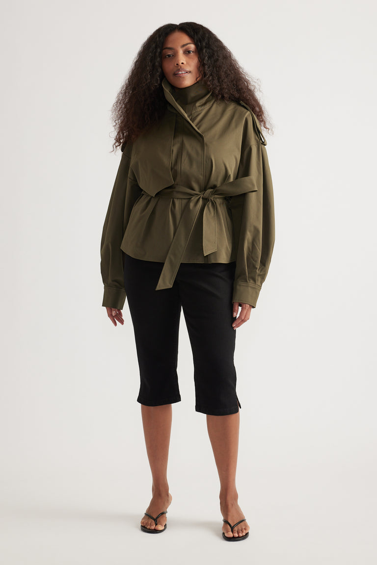 Woman wearing an olive green cropped trench coat with a tie waist, black cropped pants and thong sandals