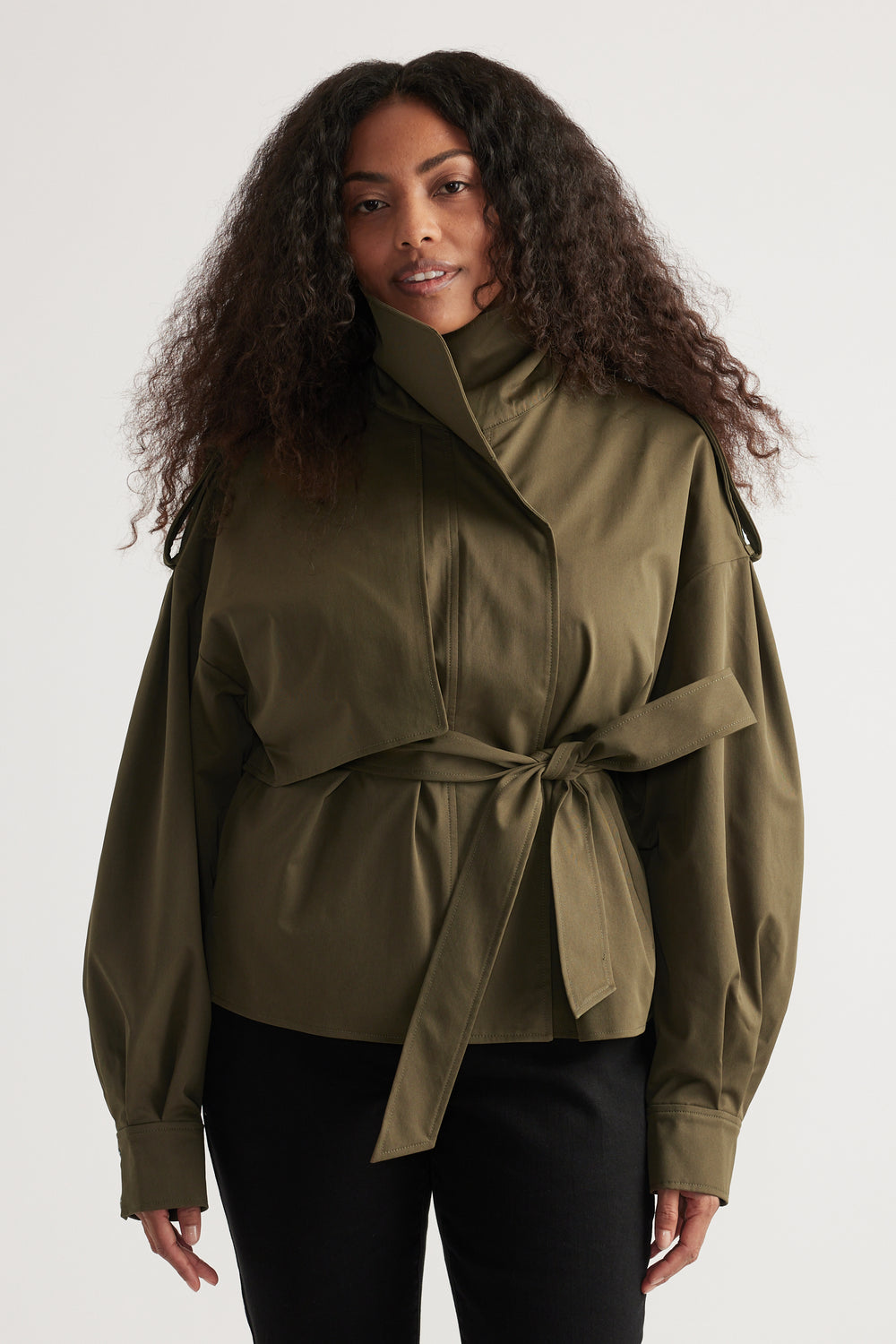 Woman wearing an olive cropped trench coat with a belted waist and black pants