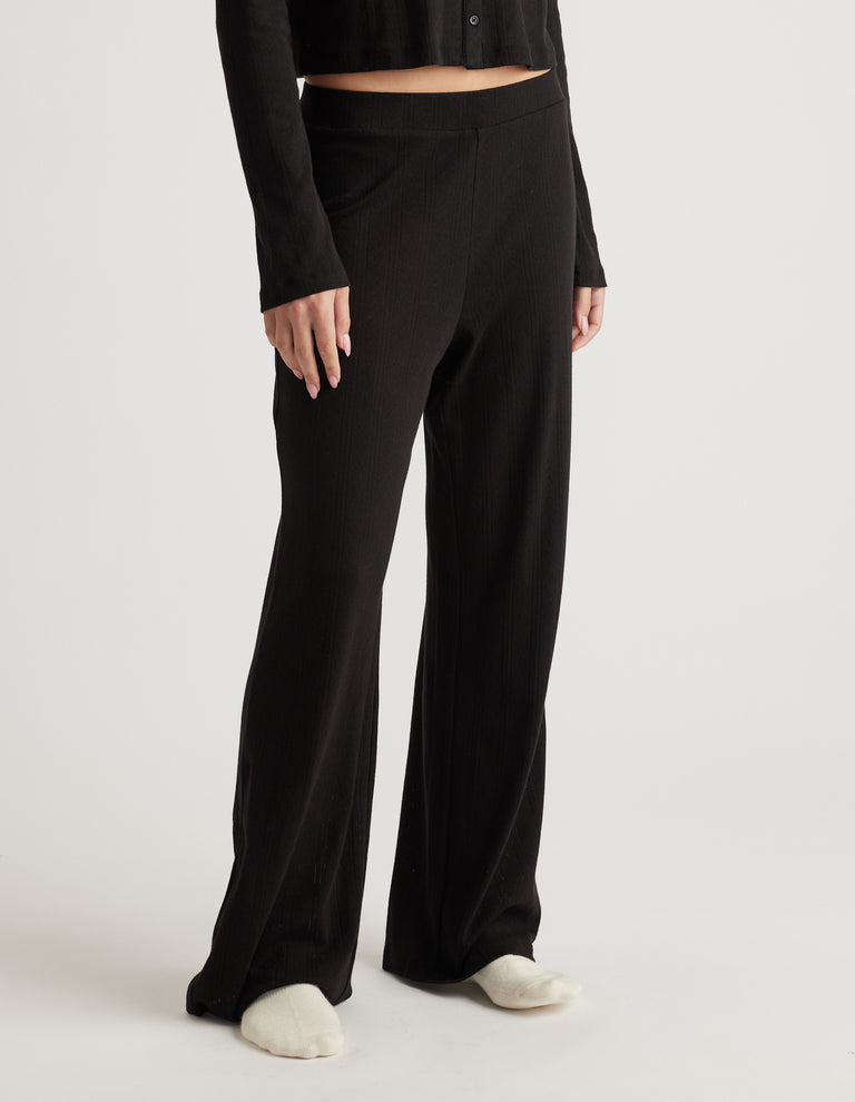 Woman wearing black wide-leg sleep pants with a subtle pointelle texture and an elastic waistband, paired with a black top